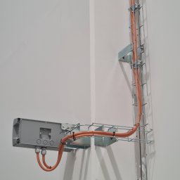 PROINSTAL - Pomarańczowe przewody elektryczne prowadzone w metalowej korytkowej trasie kablowej, przymocowanej do ściany w rogu pomieszczenia, z szarą puszką przyłączeniową.