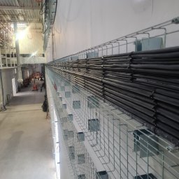 PROINSTAL - Metalowa trasa kablowa z ułożonymi czarnymi przewodami w hali przemysłowej, widok z góry, w tle widoczni pracownicy i maszyny budowlane.