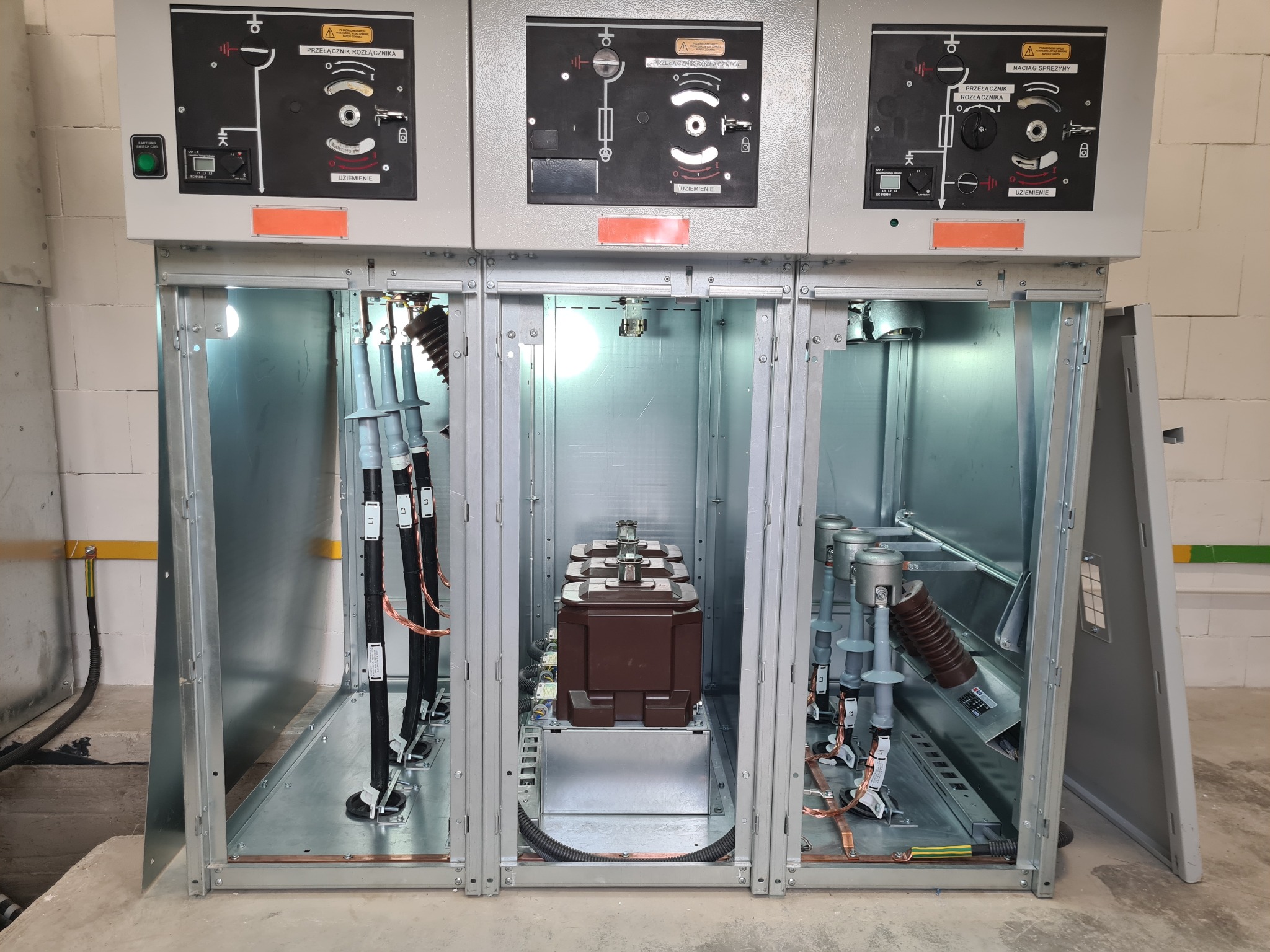 Rozdzielnica elektryczna średniego napięcia z widocznymi elementami łączeniowymi, transformatorami prądowymi i przewodami, wewnątrz metalowej obudowy z otwartymi drzwiami.