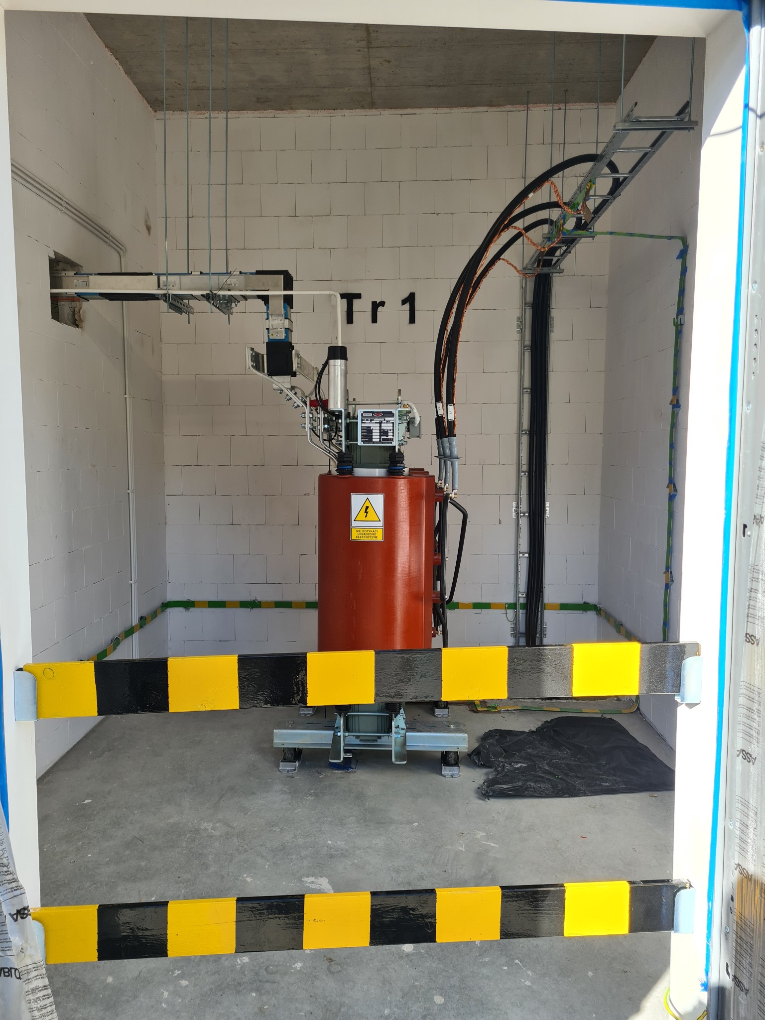 Pomarańczowy transformator w białym pomieszczeniu z betonowym sufitem, zabezpieczony barierą w żółto-czarne pasy, kable prowadzone po drabinkach kablowych.