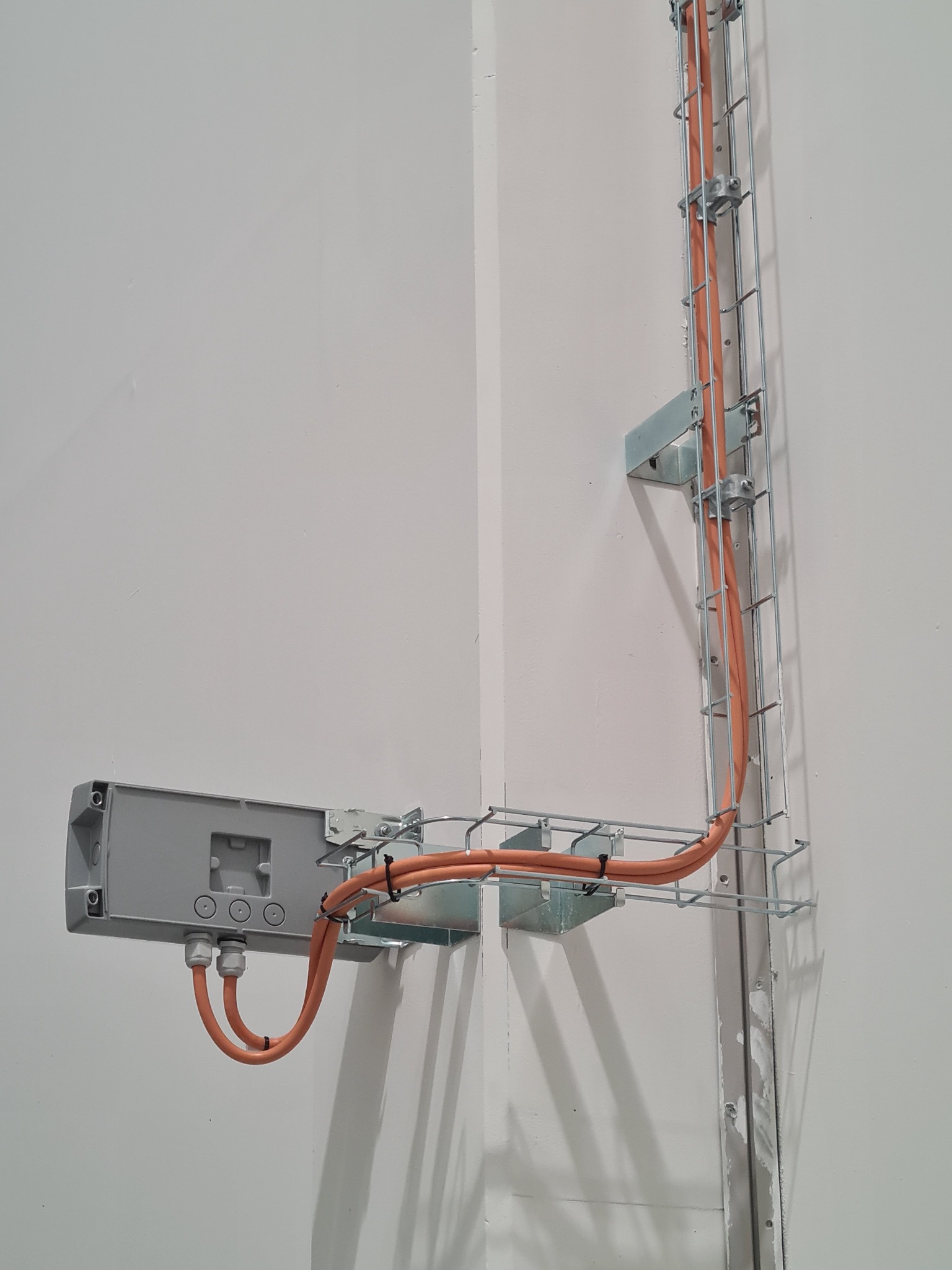 Pomarańczowe przewody elektryczne prowadzone w metalowej korytkowej trasie kablowej, przymocowanej do ściany w rogu pomieszczenia, z szarą puszką przyłączeniową.