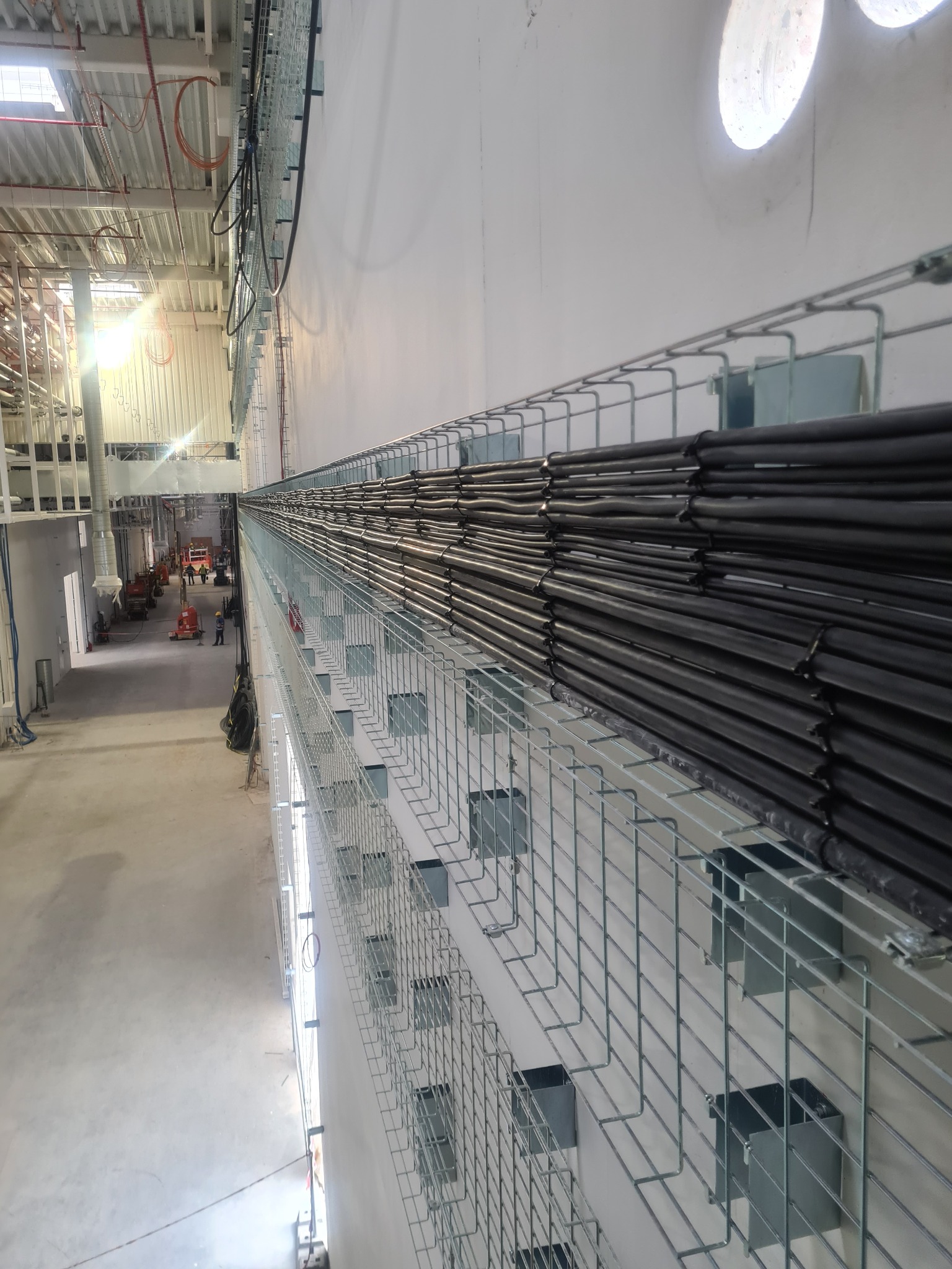 Metalowa trasa kablowa z ułożonymi czarnymi przewodami w hali przemysłowej, widok z góry, w tle widoczni pracownicy i maszyny budowlane.
