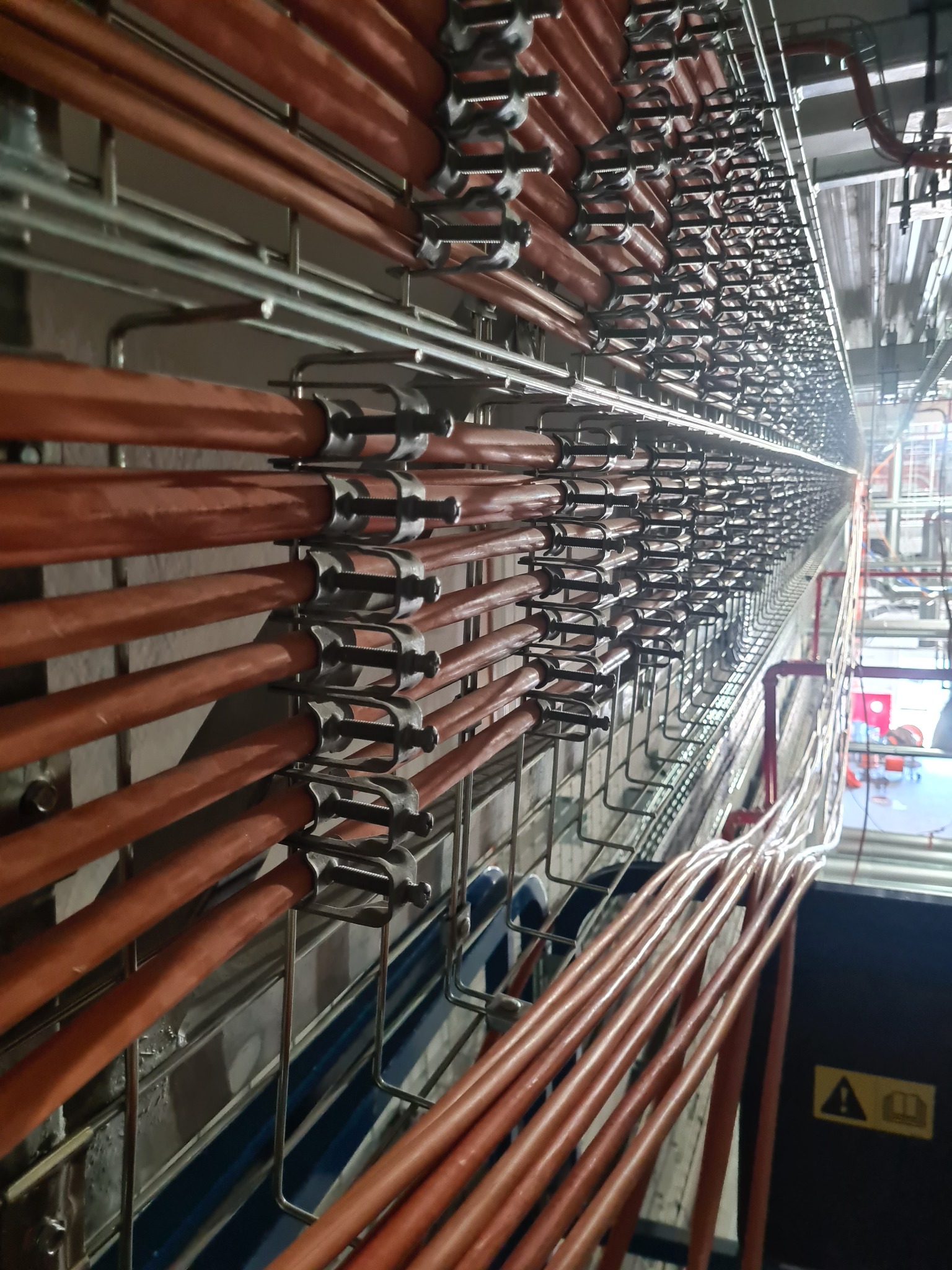 Rzędy pomarańczowych kabli ułożone w metalowych korytach, mocowane uchwytami, widok z dołu w kierunku oddalającego się tunelu instalacyjnego.
