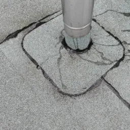 Szary dach pokryty papą z widocznym kominem i śladami naprawy uszczelniającej wokół niego.