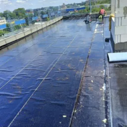 Prace dekarskie na dachu płaskim: układanie hydroizolacji z papy termozgrzewalnej, widoczny pracownik z odkurzaczem przemysłowym, zabezpieczenia krawędzi dachu, budynki w tle.