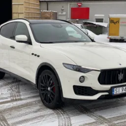 Biały samochód Maserati Levante z czarnymi felgami i przyciemnionymi szybami, zaparkowany na śniegu przed budynkiem magazynowym.