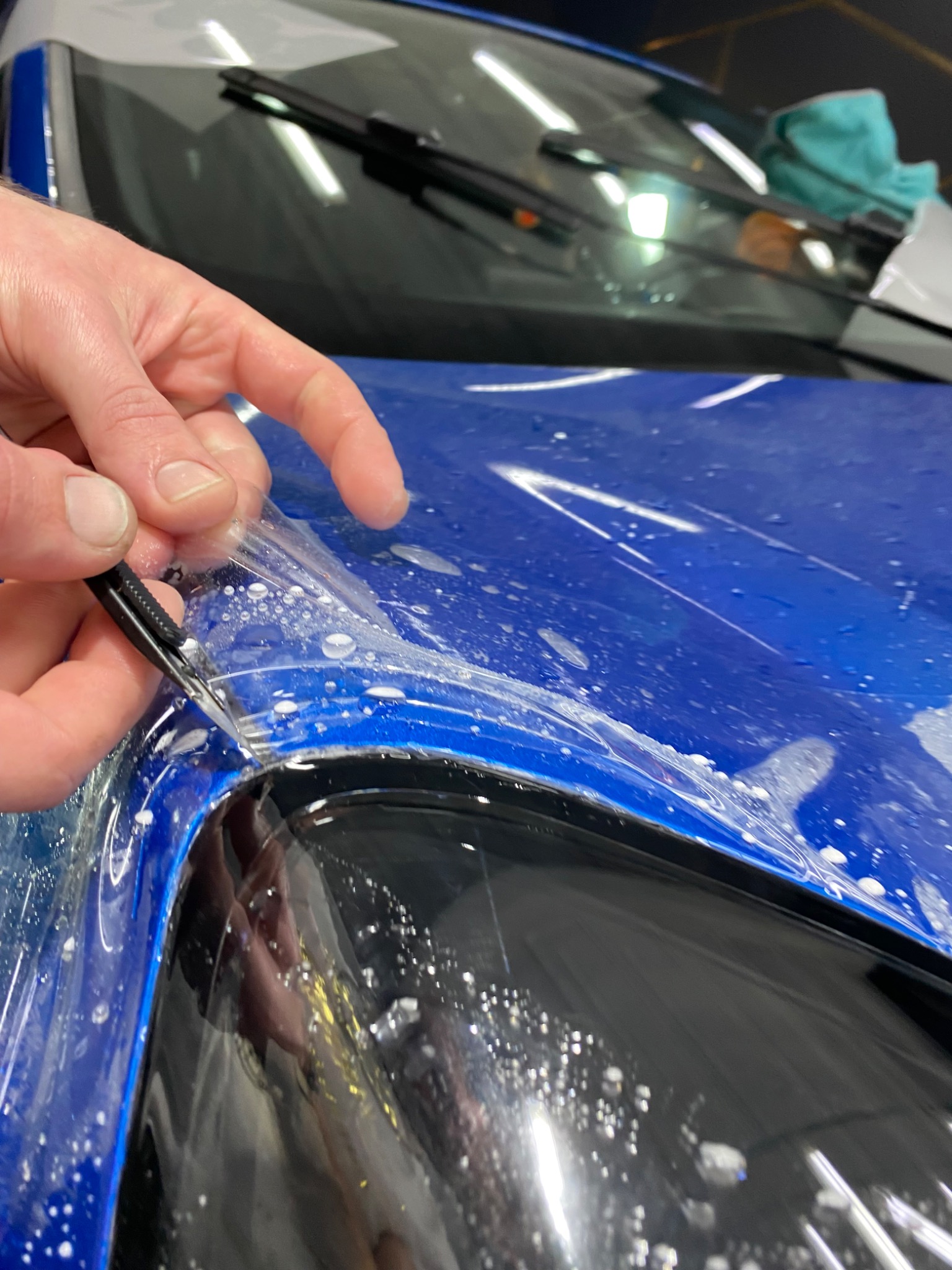 Aplikacja folii ochronnej na niebieski samochód w Czeladzi, widoczne narzędzie i dłoń podczas usuwania nadmiaru folii, krople wody na powierzchni.