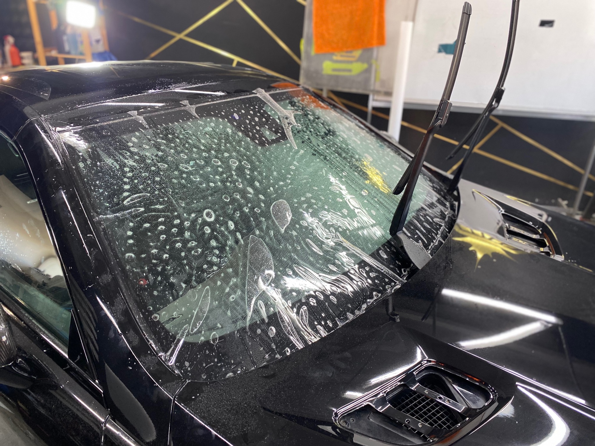 Przygotowanie do przyciemniania szyby przedniej czarnego samochodu, widoczne bąbelki wody pod folią ochronną.