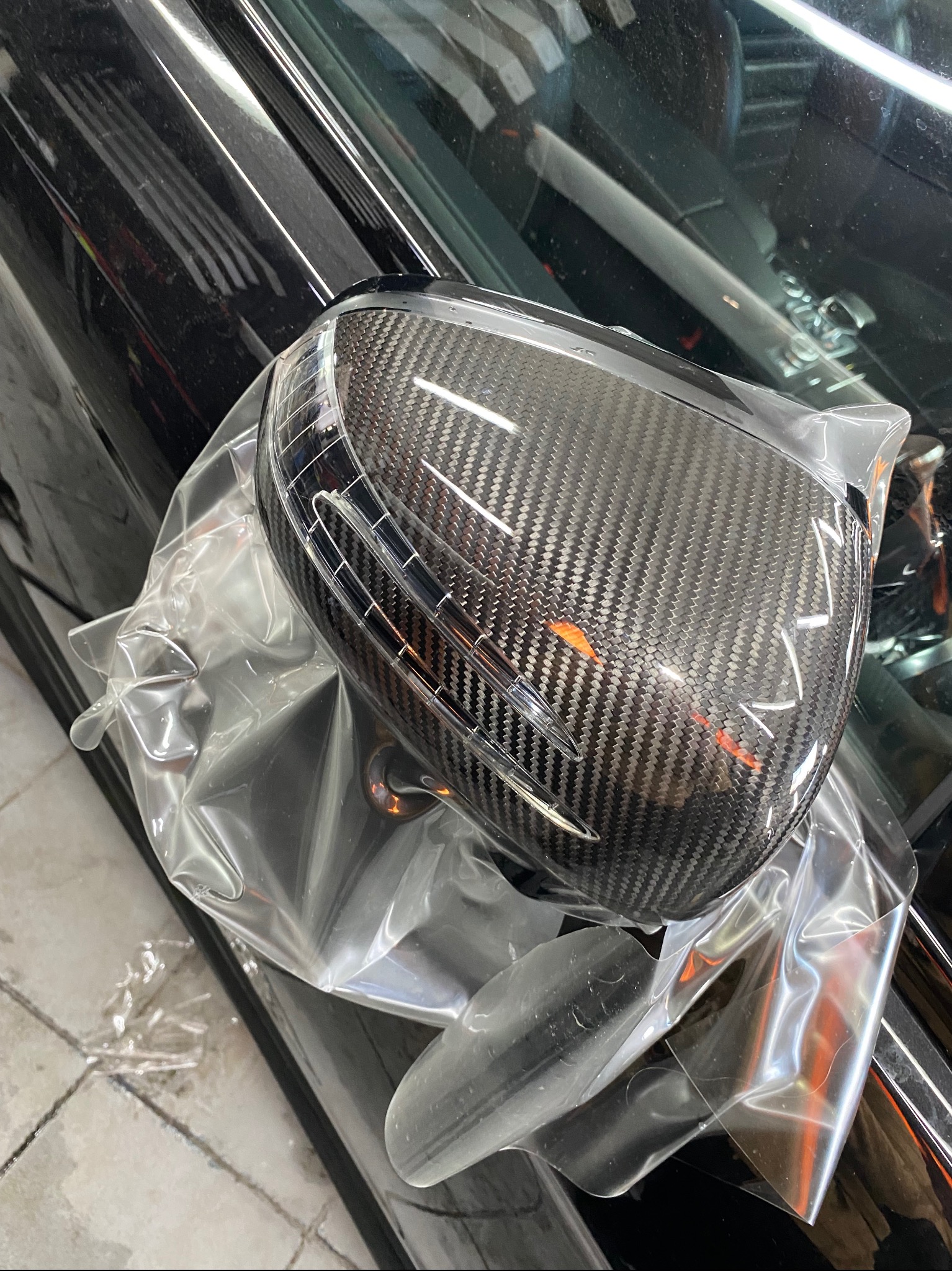 Lusterko samochodowe pokryte folią karbonową, jeszcze w folii ochronnej, zamontowane na czarnym samochodzie. Widoczny wzór karbonu i odbicia światła na folii.