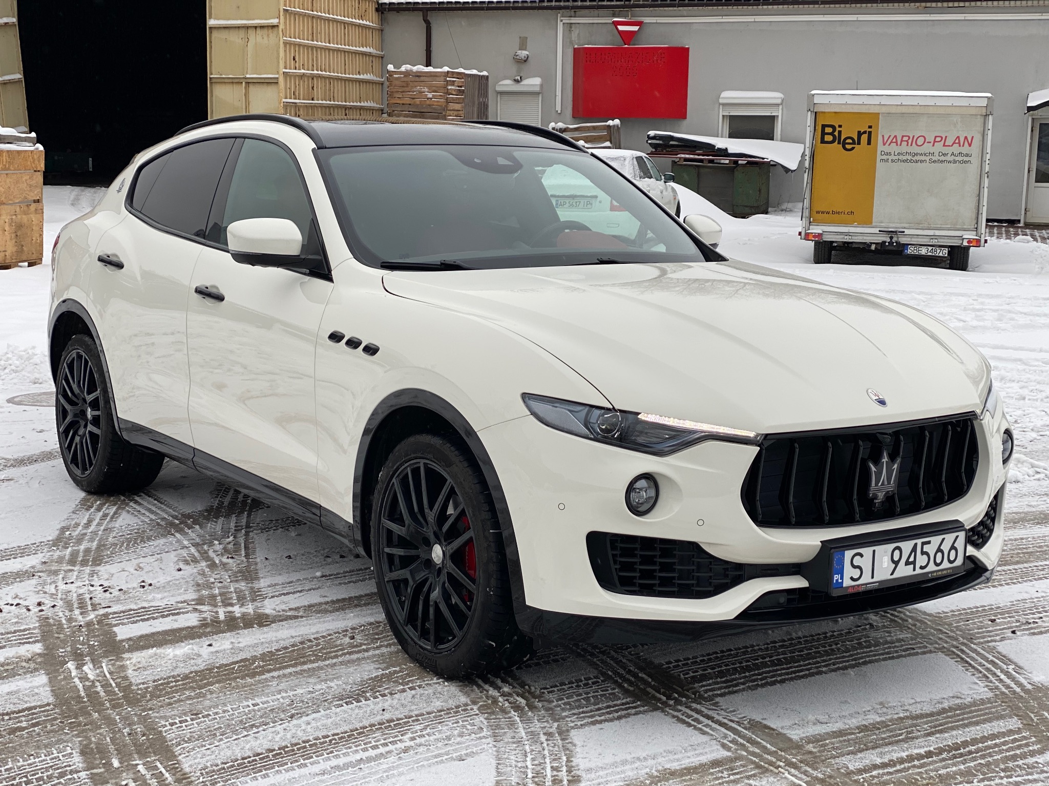 Biały samochód Maserati Levante z czarnymi felgami i przyciemnionymi szybami, zaparkowany na śniegu przed budynkiem magazynowym.