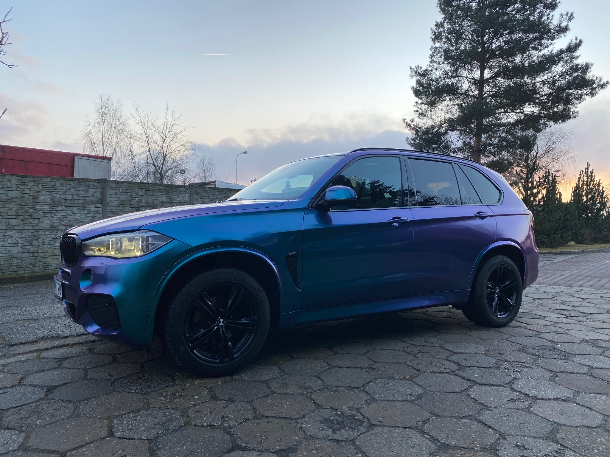 BMW X5 z efektem kameleona na karoserii, zmieniającym kolor od turkusowego do fioletowego, zaparkowany na kostce brukowej, z widocznym tłem drzew i zachodzącego słońca.