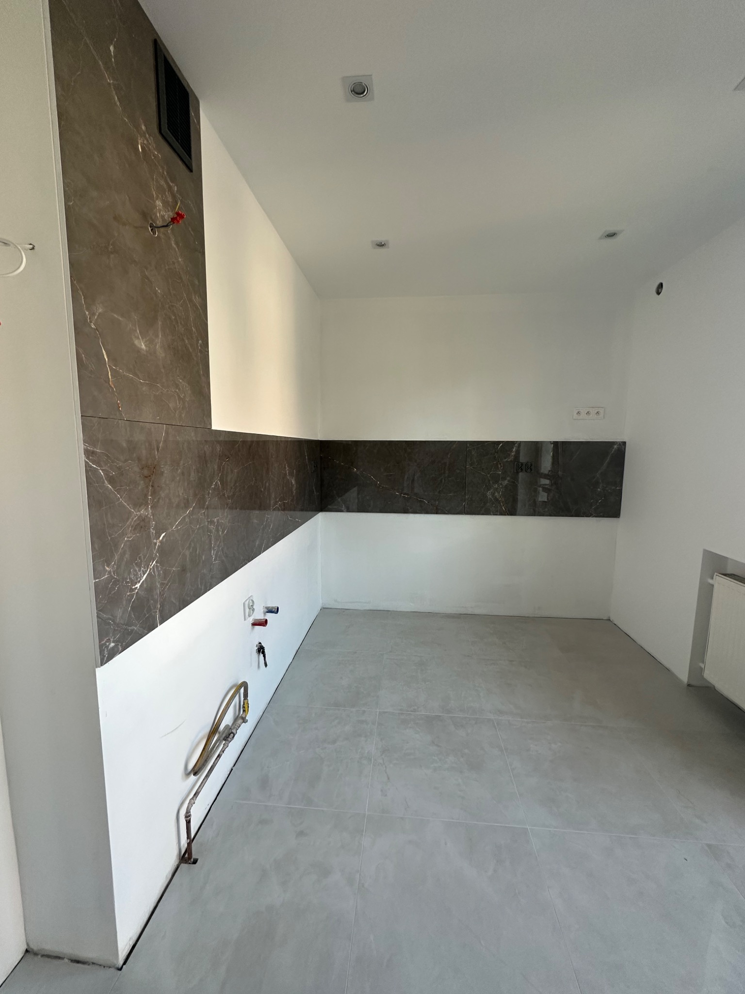 Wnętrze kuchni w trakcie remontu, widoczne płytki imitujące marmur na ścianach, szare płytki podłogowe i wystające rury instalacji wodnej.