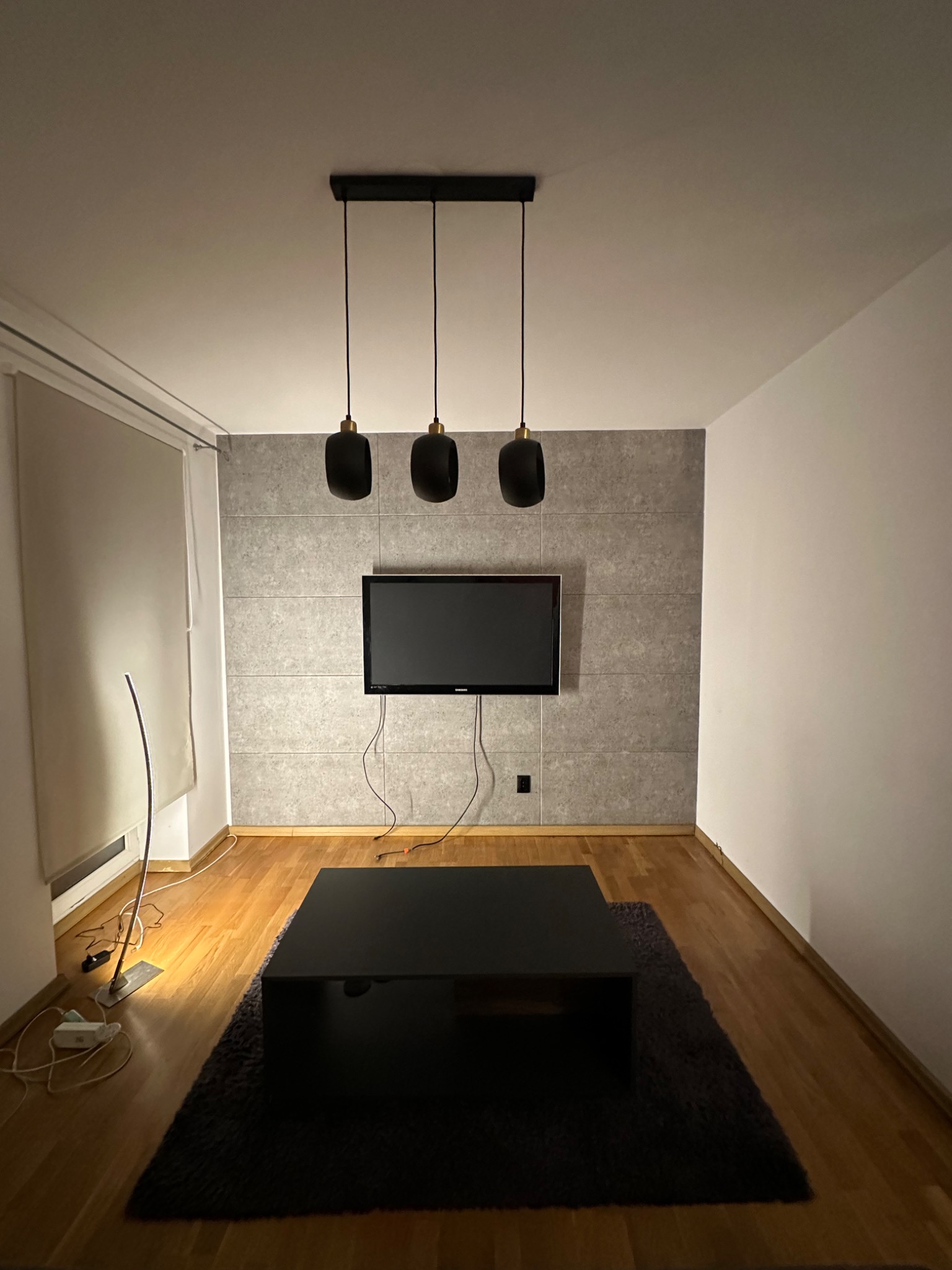 Nowoczesny salon z szarą ścianą z płytek imitujących kamień, wiszącym telewizorem, czarnym stolikiem kawowym i trzema czarnymi lampami wiszącymi, oświetlonymi przez lampę podłogową.