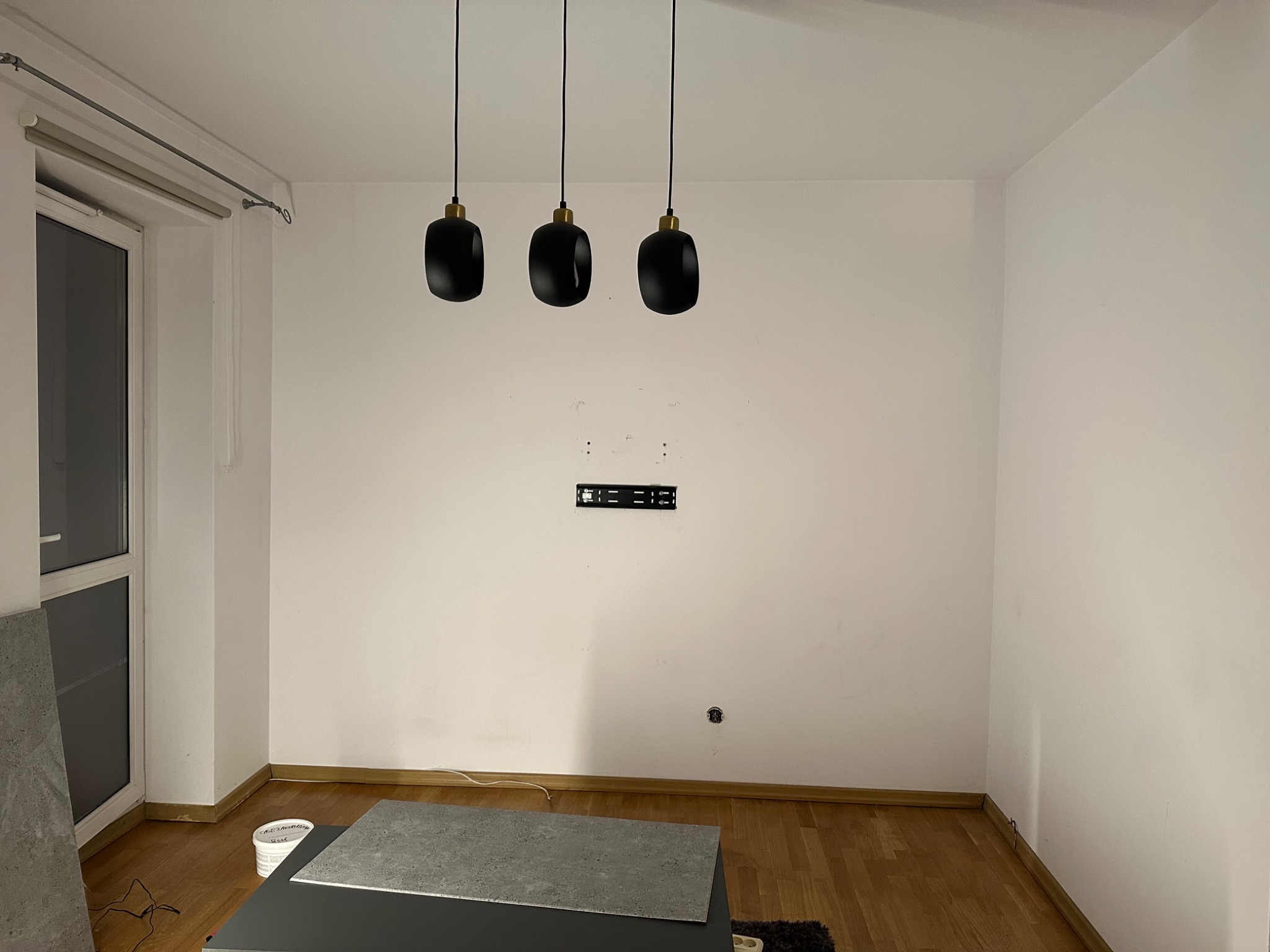 Półsurowy pokój z trzema czarnymi lampami wiszącymi, płytą montażową na ścianie, przygotowanymi płytami podłogowymi i niedoskonałościami na ścianie, ukazujący etap prac wykończeniowych.
