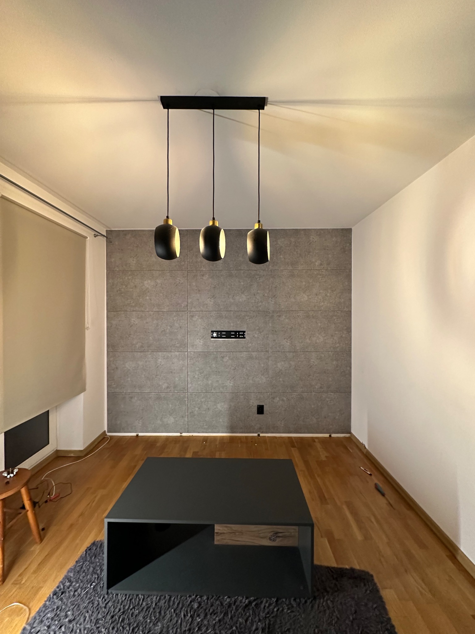 Salon po remoncie: ściana wyłożona szarymi panelami imitującymi beton, podwieszana lampa z trzema czarnymi kloszami, drewniana podłoga, szary dywan i nowoczesny stolik kawowy.