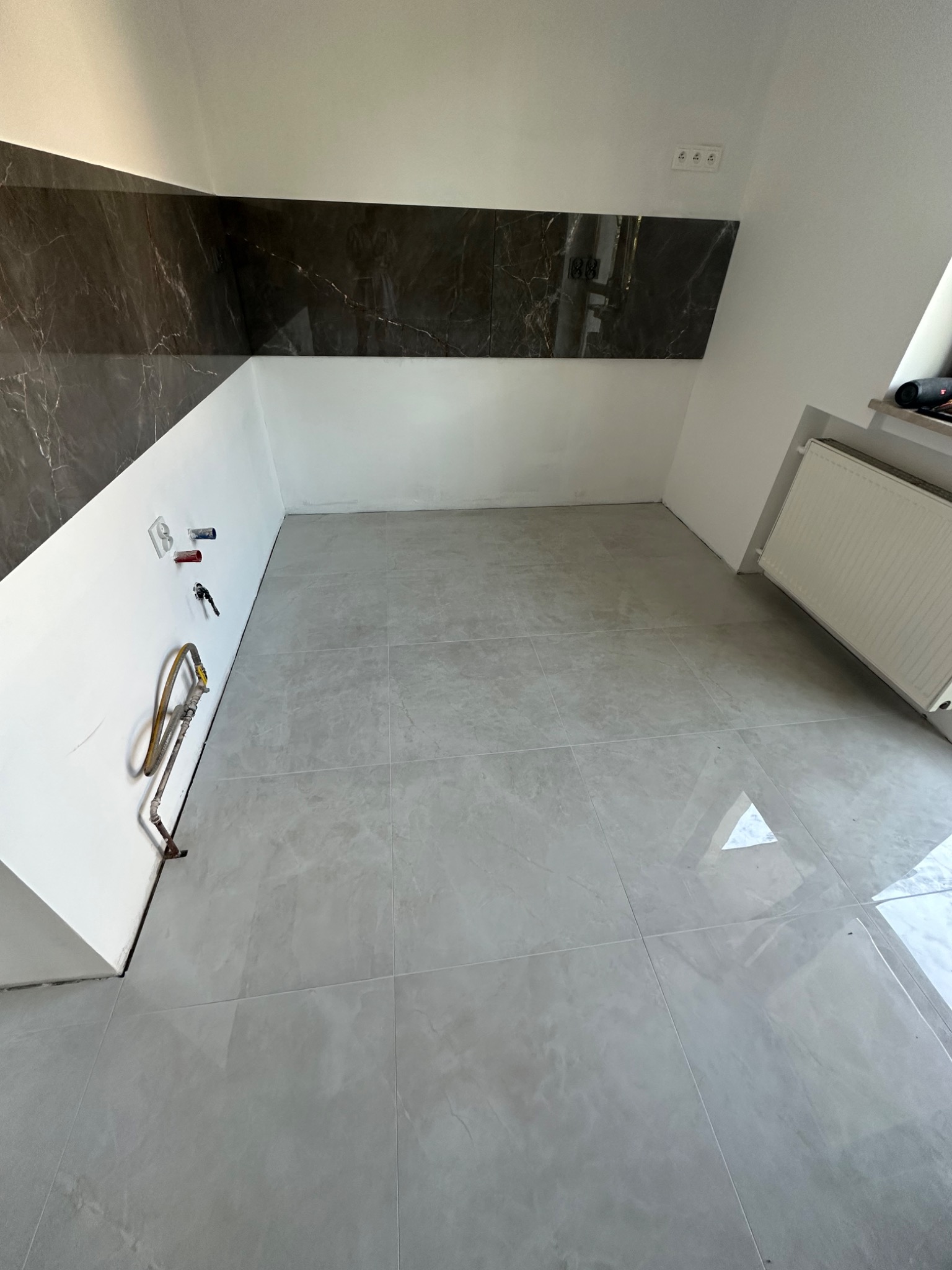 Świeżo położone, błyszczące, szare płytki podłogowe i częściowo ułożone, ciemne płytki imitujące marmur na ścianie w trakcie prac wykończeniowych, widoczne wystające rury i gniazdka elektryczne.