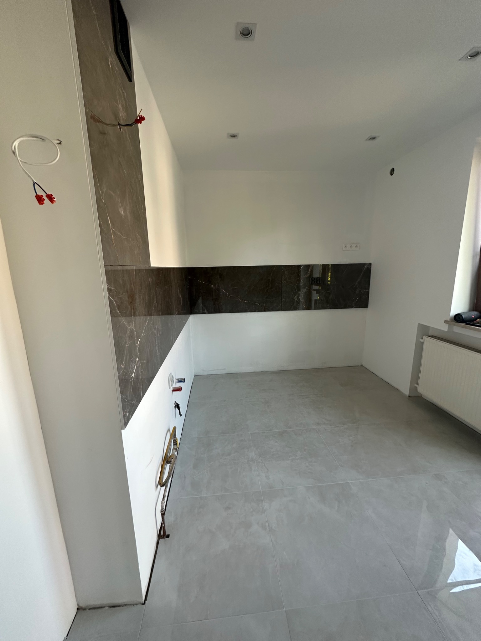 Wnętrze łazienki w trakcie remontu: szare płytki podłogowe, częściowo wykończona ściana z panelami imitującymi marmur, widoczne przewody elektryczne i narzędzia instalacyjne.