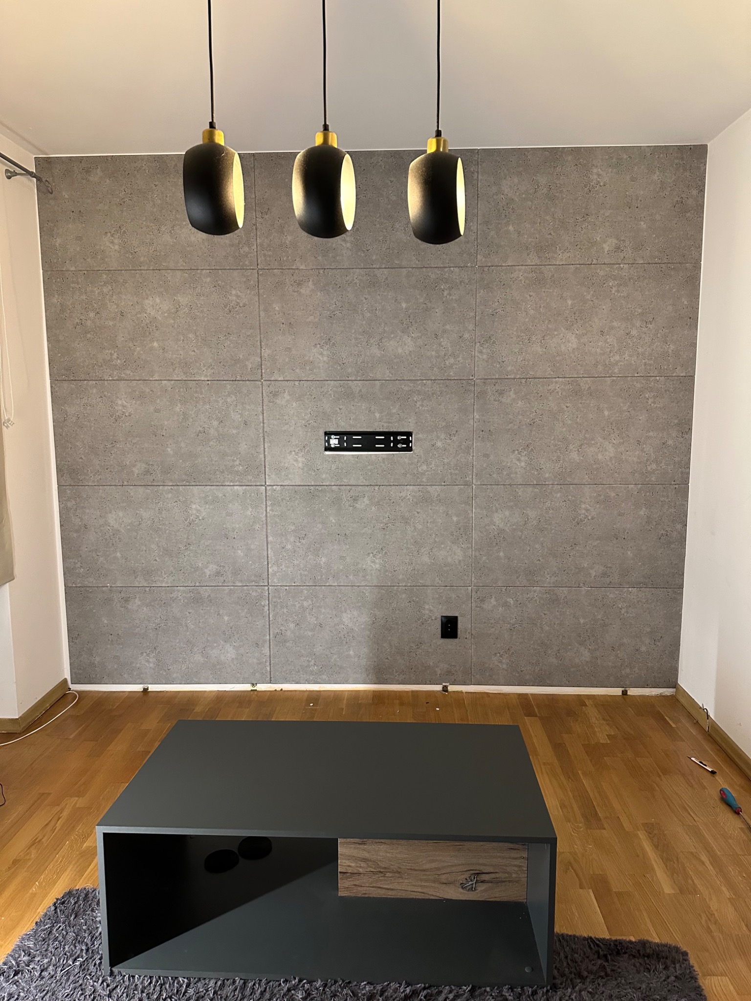 Ściana w salonie wykończona panelami imitującymi beton, z zamontowanym uchwytem na telewizor i gniazdkiem elektrycznym, oświetlona trzema wiszącymi lampami z czarnymi kloszami i złotymi elementami,...