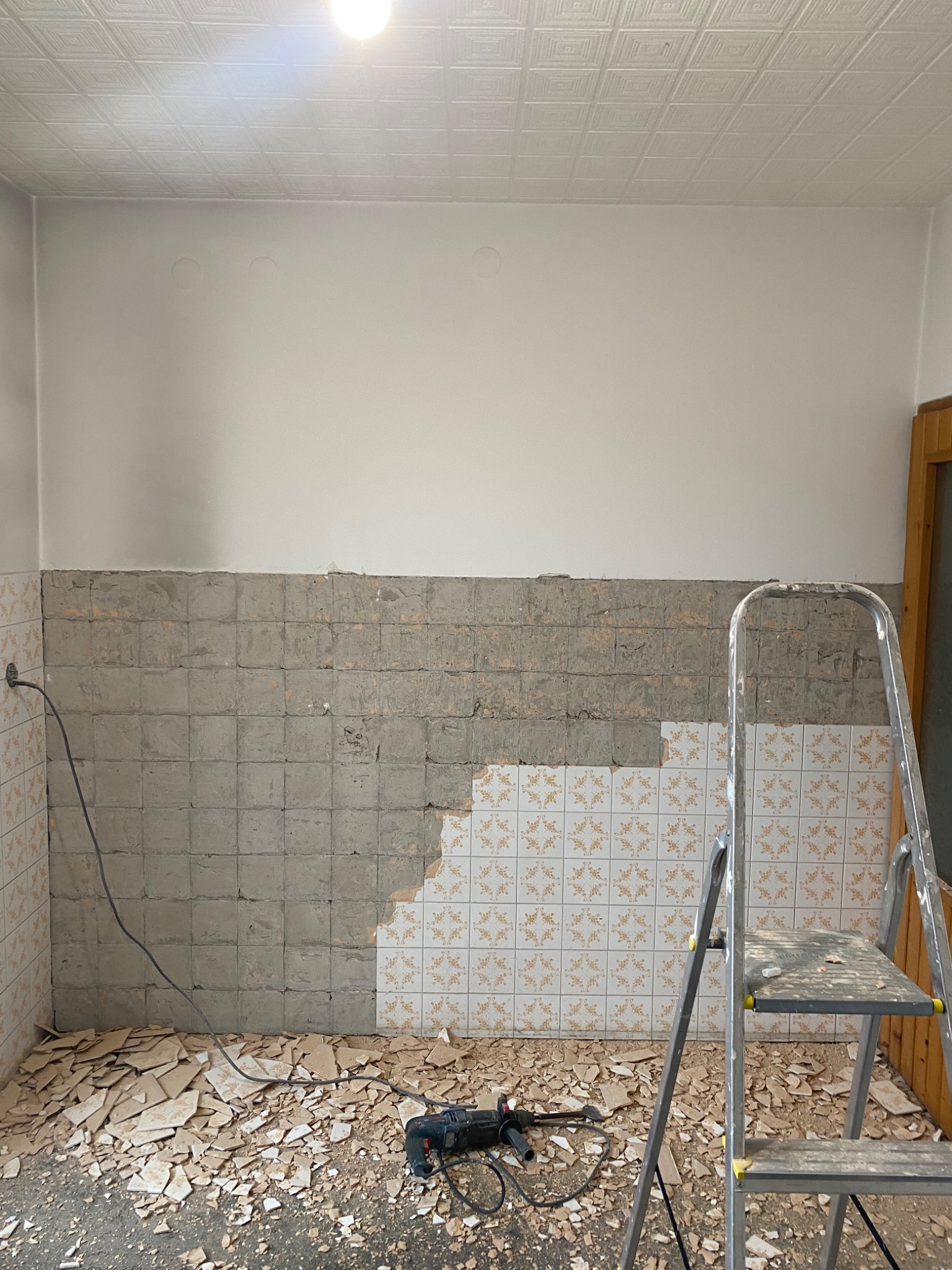 Demontaż płytek ściennych w kuchni, widoczny fragment ściany z resztkami kleju, gruz na podłodze, drabina i wiertarka udarowa.