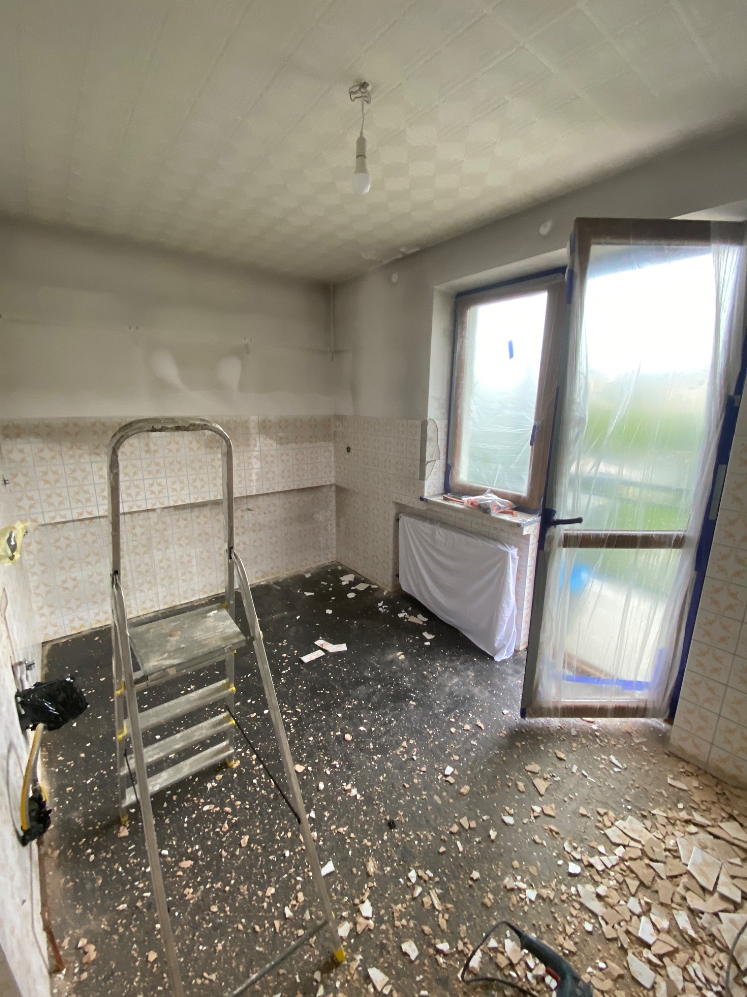Kuchnia w trakcie remontu: stara glazura częściowo usunięta, drabina aluminiowa, okno zabezpieczone folią, gruz na podłodze, widoczna instalacja elektryczna.