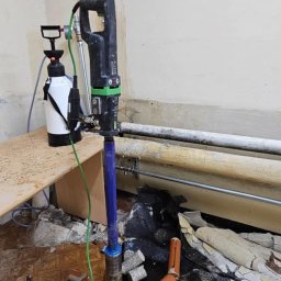 M.S INSTAL-MAT Usługi Hydrauliczne - Wiertnica rdzeniowa z osprzętem na tle ściany z rurami i wywierconymi otworami. Widoczne fragmenty gruzu i starej podłogi. Praca w trakcie wiercenia otworów.