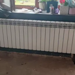 Nowy, biały grzejnik aluminiowy zamontowany przy oknie na tle ciemnej ściany. Widoczne ślady prac remontowych na podłodze i ścianie.