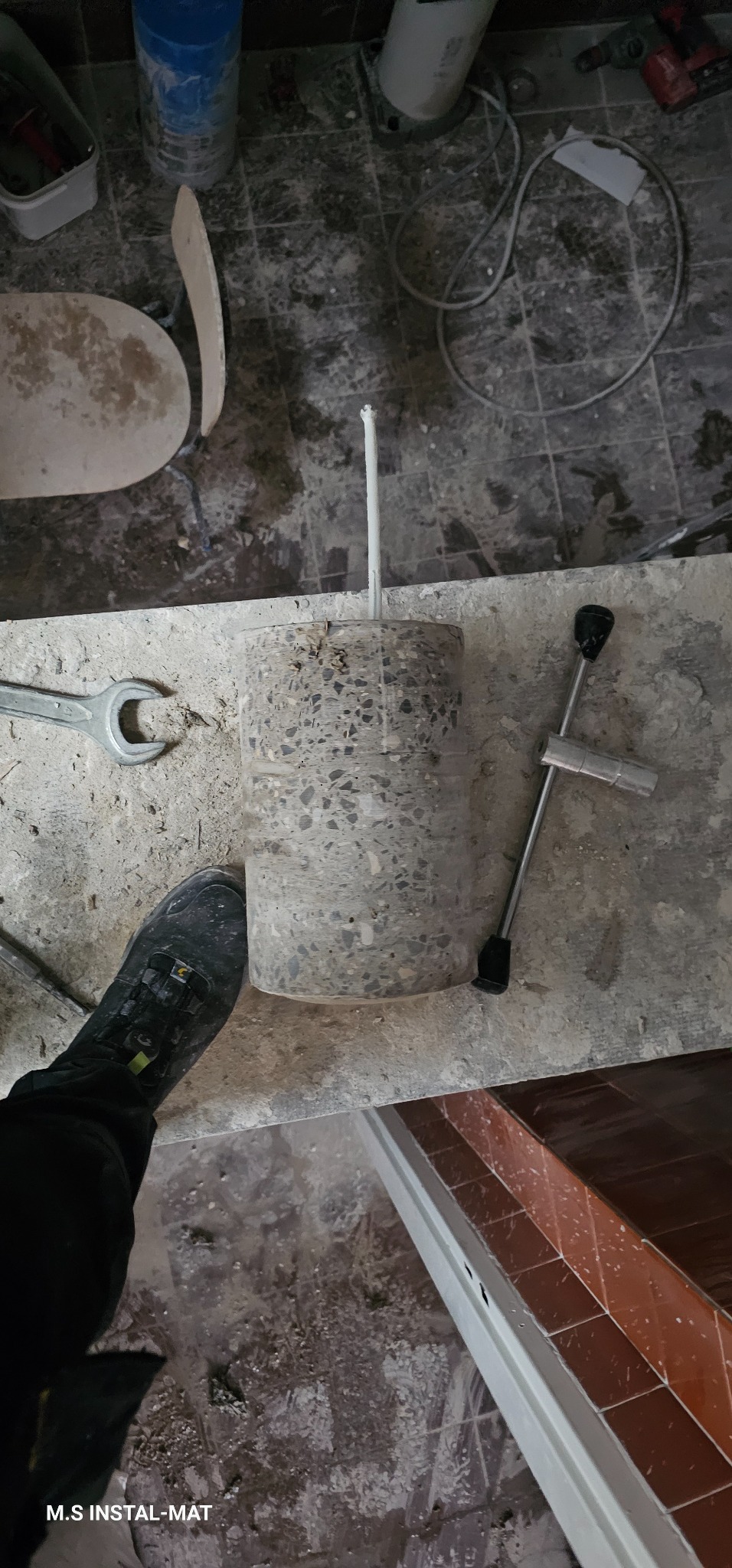 Rdzeń betonowy z wystającą rurką na tle remontowanego pomieszczenia. Widoczne narzędzia hydrauliczne i fragment buta. Szare płytki podłogowe.