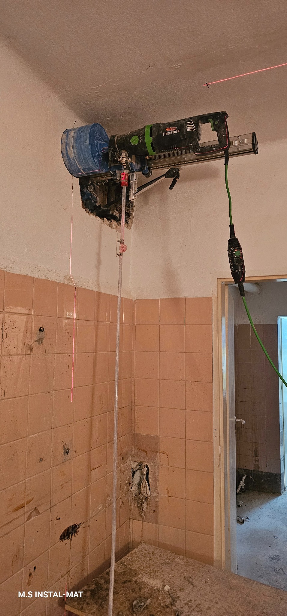 Wiertnica Eibenstock w trakcie pracy, wiercenie otworów w ścianie z różowych płytek. Widoczna wiązka lasera wyznaczająca linię wiercenia. W tle otwarte drzwi.