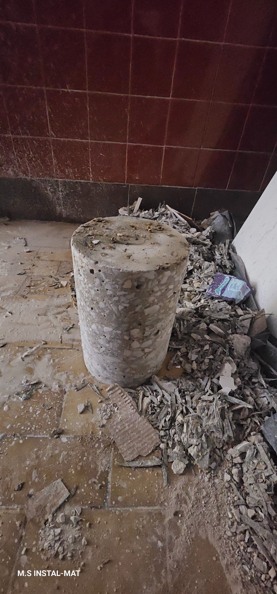 Rdzeń betonowy po wierceniu w podłodze, otoczony gruzem i fragmentami płytek. Widoczne ślady wiercenia i uszkodzenia podłogi. Tło stanowią brudne, brązowe płytki ścienne.