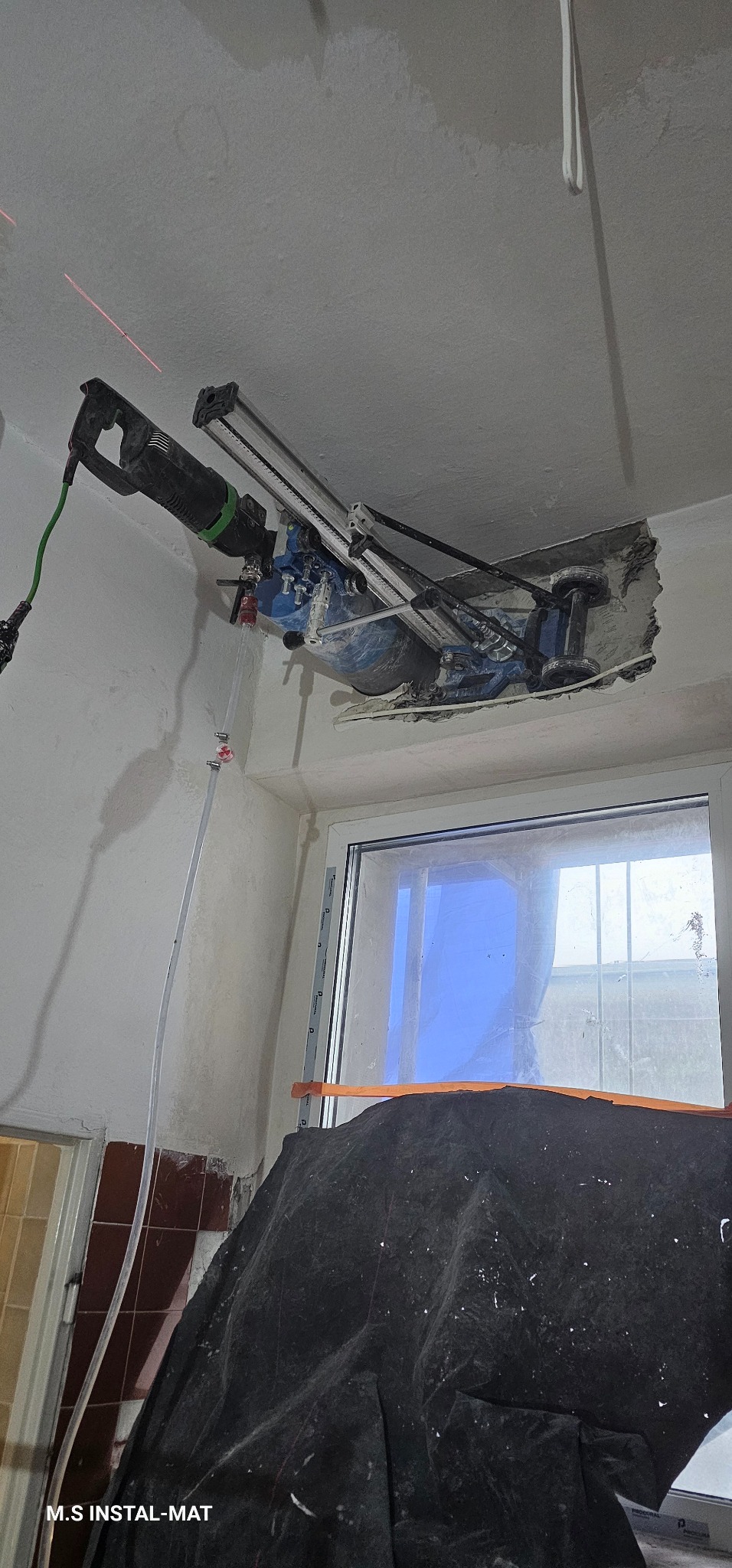 Wiertnica diamentowa mocowana do sufitu, wiercąca otwór. Widoczny laser wyznaczający linię cięcia i osłona okna. Praca w trakcie wiercenia.