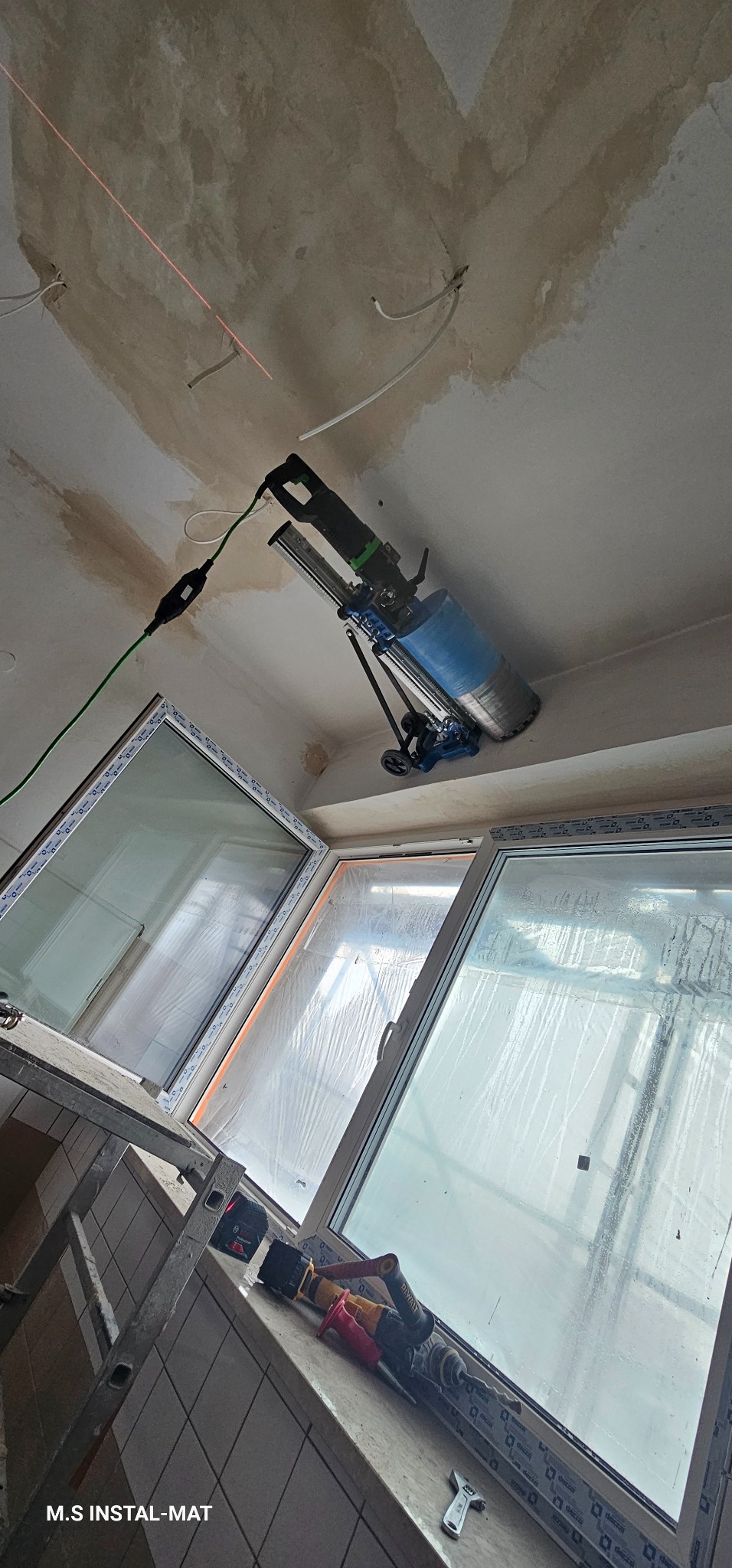 Wiercenie rdzeniowe w suficie za pomocą wiertnicy diamentowej. Widoczne okna dachowe, drabina i narzędzia. Czerwona linia lasera wyznacza kierunek wiercenia.