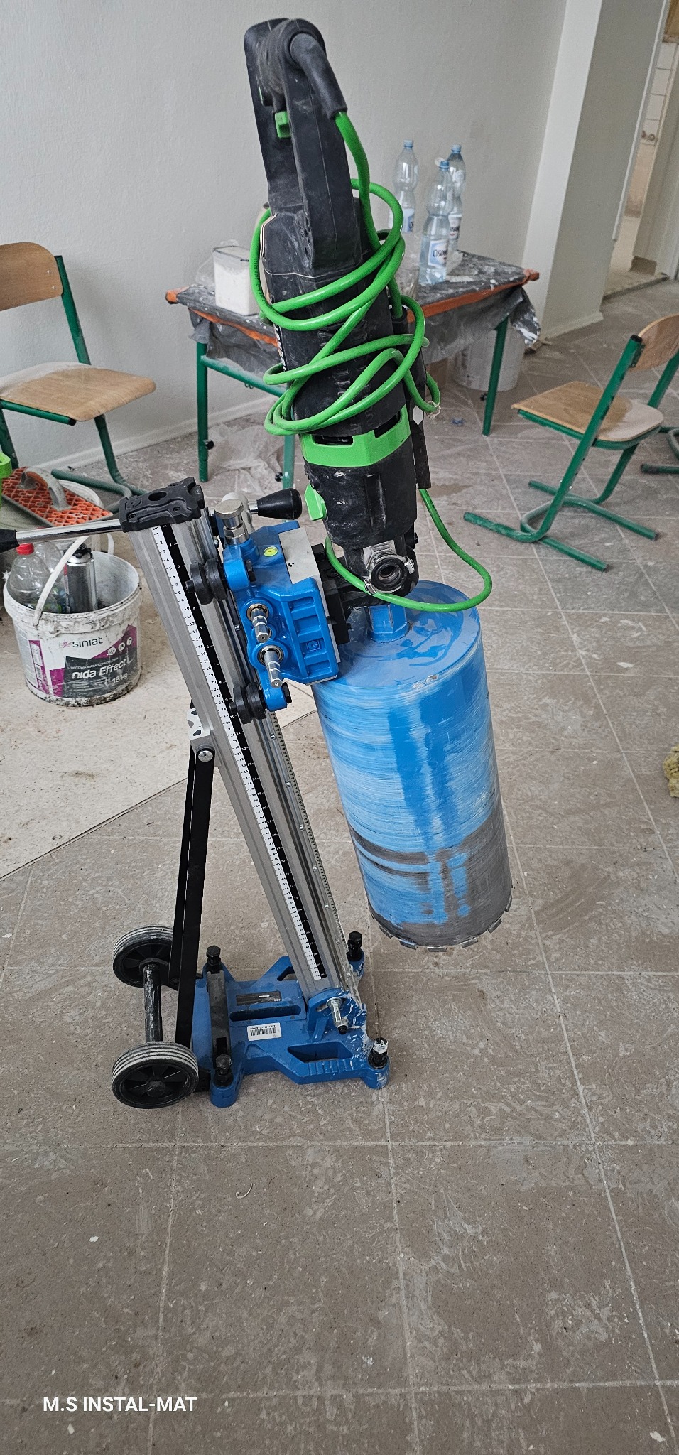 Wiertnica diamentowa z wiertłem koronowym na posadzce z płytek. Widoczny przewód zasilający i elementy konstrukcyjne w kolorze niebieskim. W tle krzesła i stolik.