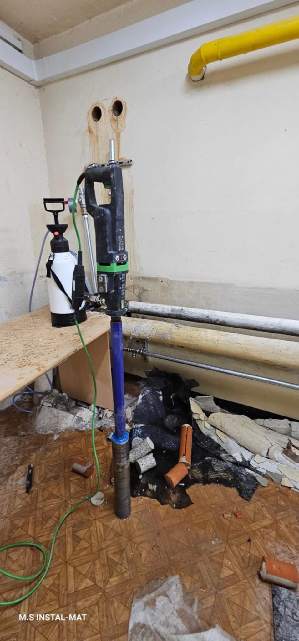 Wiertnica rdzeniowa z osprzętem na tle ściany z rurami i wywierconymi otworami. Widoczne fragmenty gruzu i starej podłogi. Praca w trakcie wiercenia otworów.