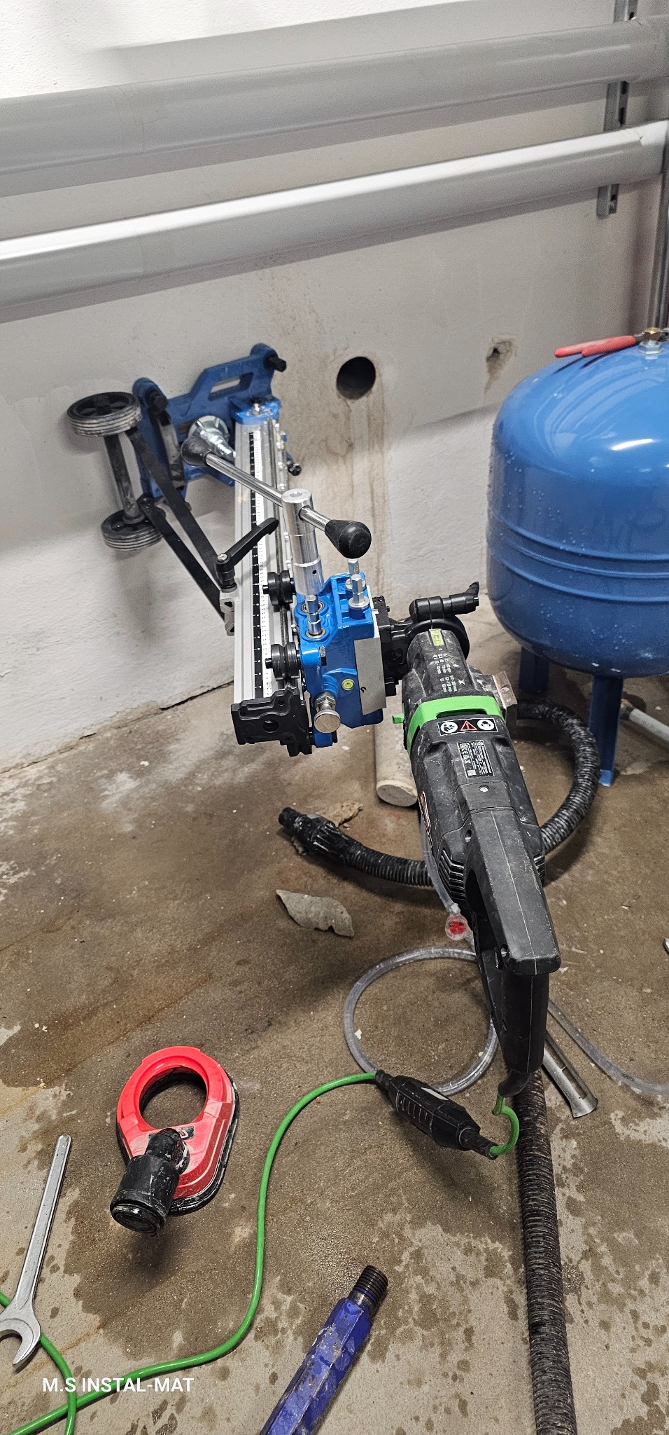 Wiertnica diamentowa zamocowana do ściany, obok niebieski zbiornik ciśnieniowy. Widoczne narzędzia hydrauliczne i ślady wiercenia w ścianie.