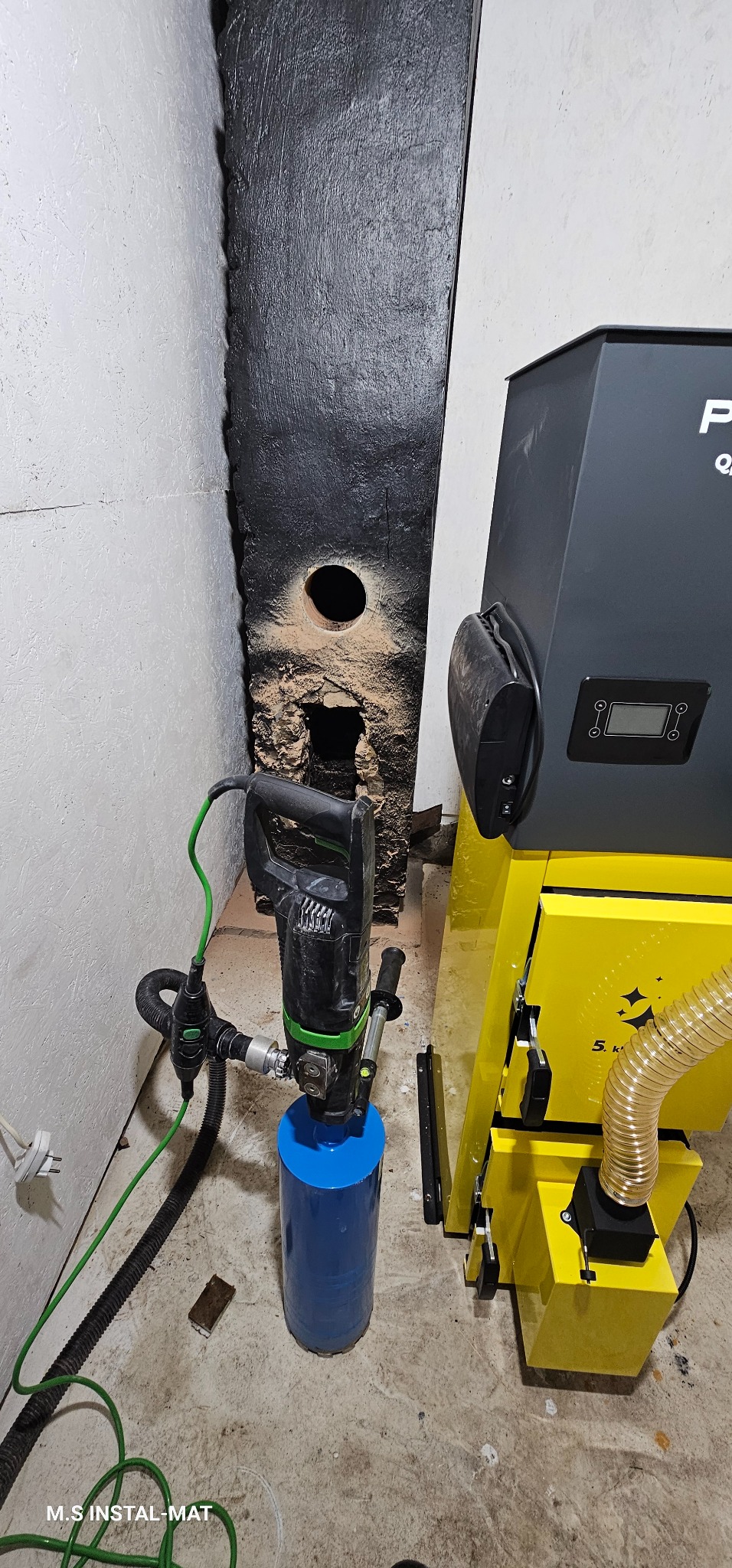 Wiertnica diamentowa z wiertłem koronowym, obok piec CO, wiercenie otworu w ścianie. Prace instalacyjne w kotłowni. Widoczne przewody i elementy instalacji.