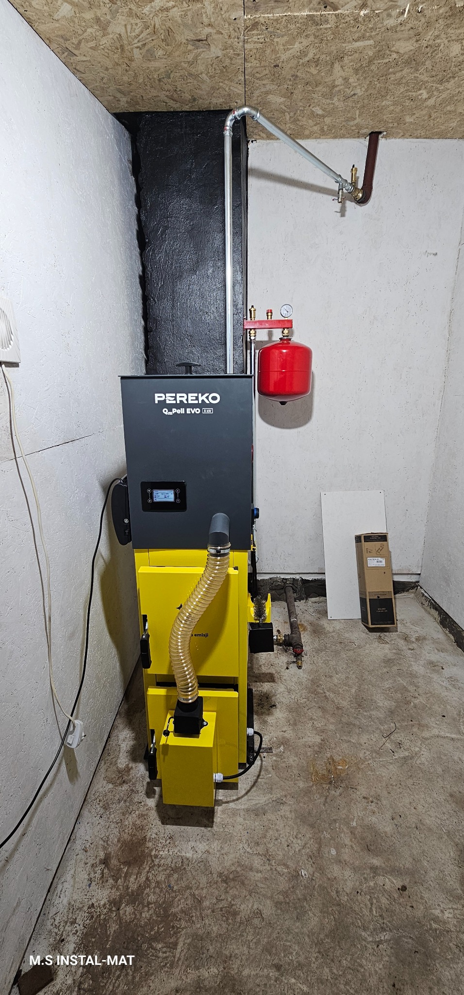 Żółto-czarny piec Pereko Qm Pell EVO 8kW z podłączoną instalacją grzewczą w kotłowni o surowych ścianach i podłodze. Widoczny manometr i rury.