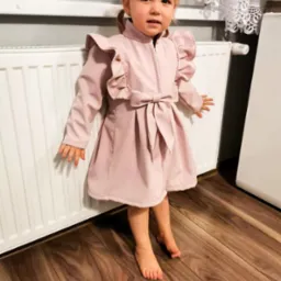 Dziewczynka w różowej sukience z falbankami i kokardą, stojąca boso na drewnianej podłodze obok białego grzejnika i okna z koronkową firanką.