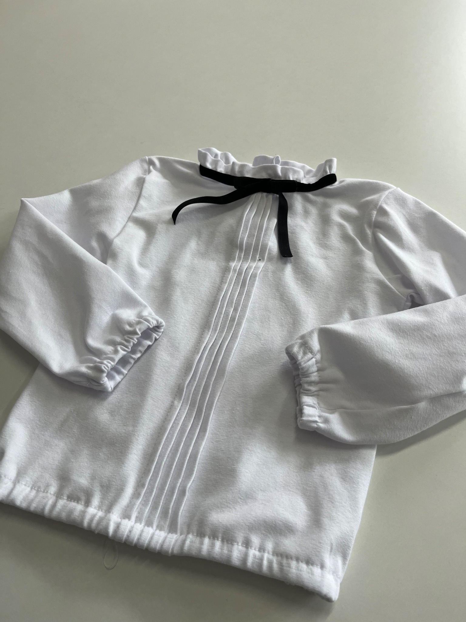 Biała bluzka dziecięca z długim rękawem, plisowanym przodem i czarną kokardką pod szyją, leżąca na białym tle.