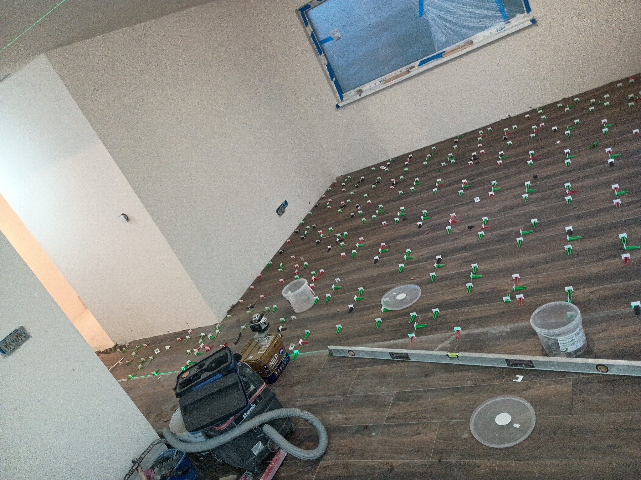 Układanie płytek imitujących drewno na podłodze z użyciem systemu poziomowania. Widoczne narzędzia glazurnicze i okno zabezpieczone folią. Praca w toku.