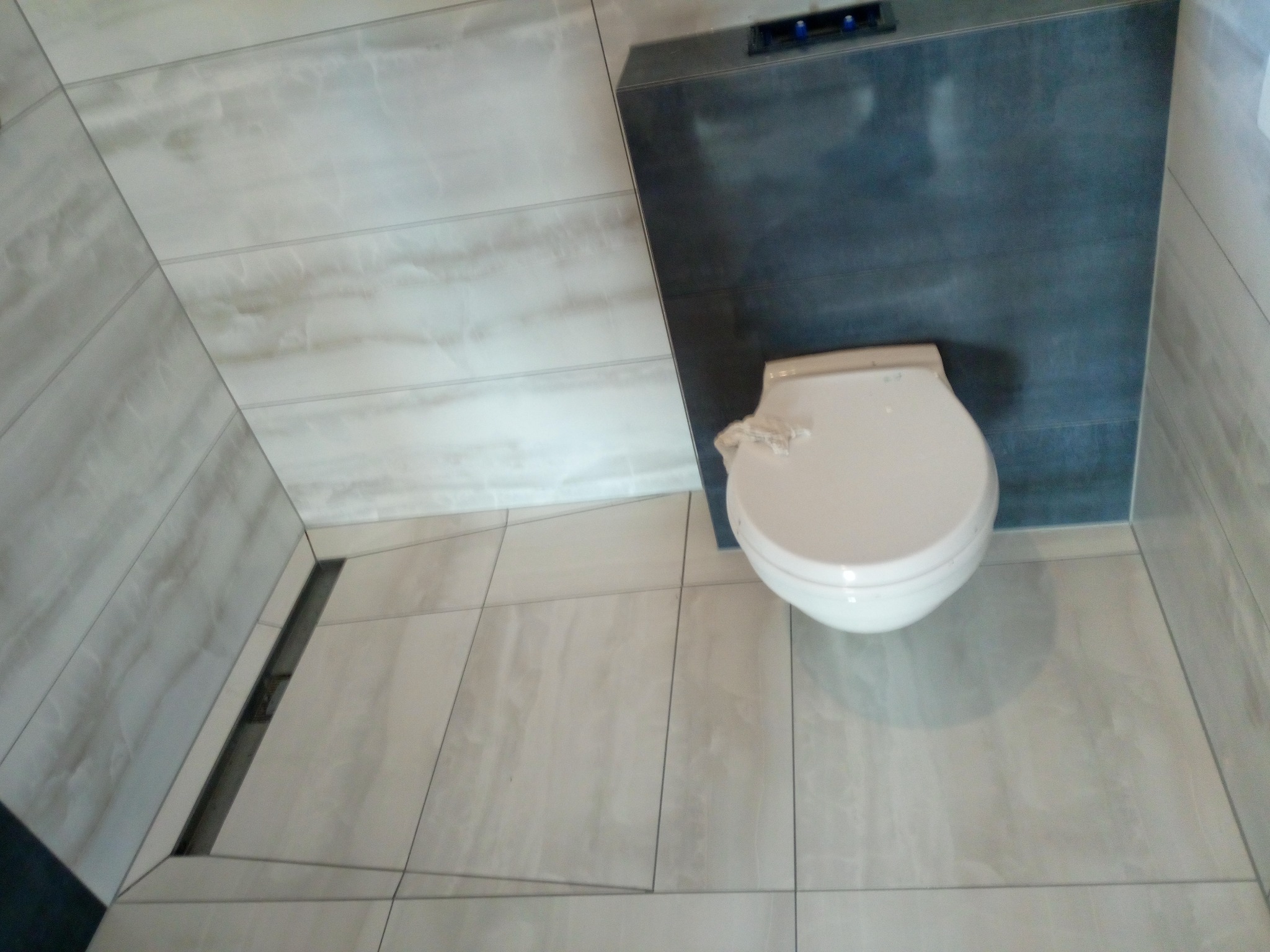 Wykończona łazienka z podwieszaną toaletą i odpływem liniowym. Jasne płytki imitujące marmur na ścianach i podłodze, ciemny zabudowany stelaż podtynkowy.