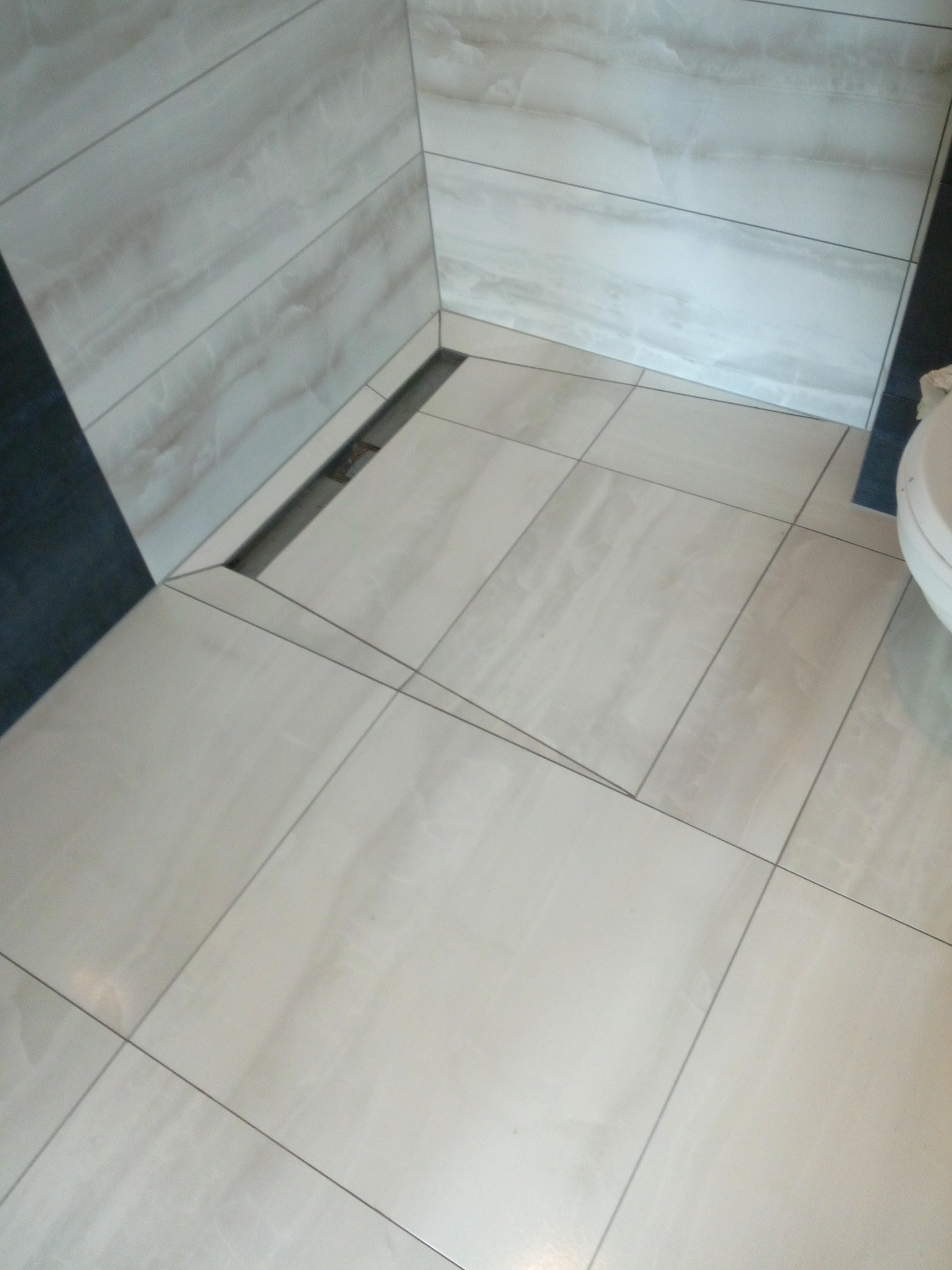 Jasne, marmurkowe płytki podłogowe i ścienne w łazience z odpływem liniowym. Minimalistyczny design, widok z góry, fragment toalety po prawej.