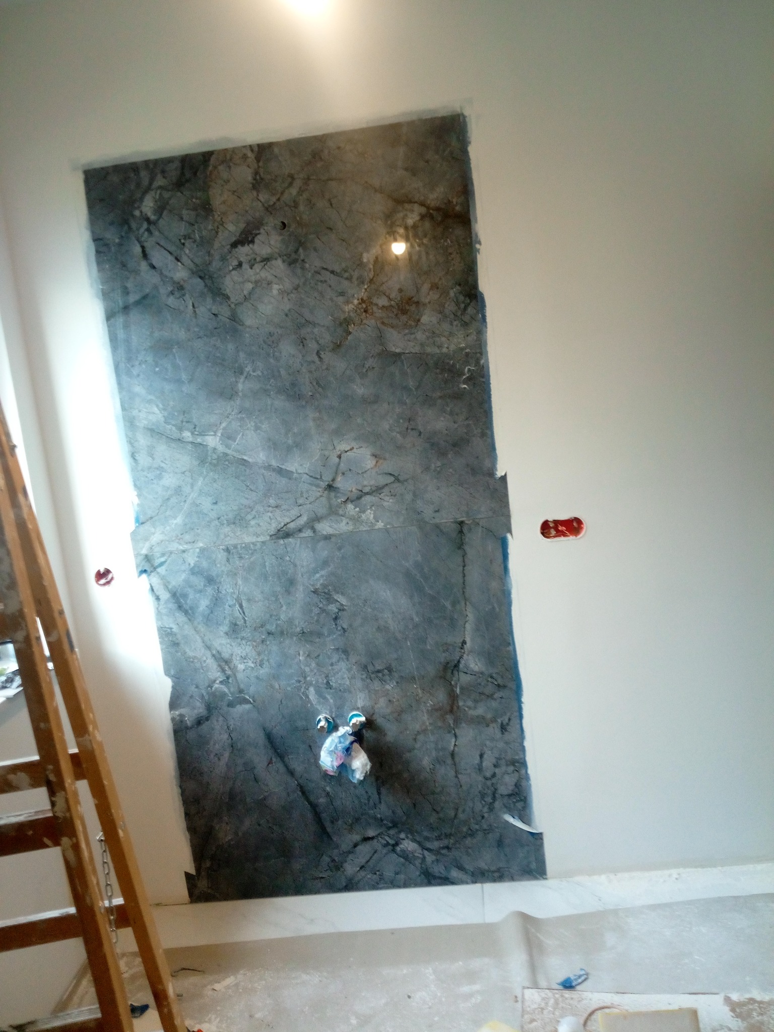 Montaż płyty imitującej marmur na ścianie w łazience. Widoczne gniazdka elektryczne, drabina malarska i fragment podłogi. Prace wykończeniowe w toku.