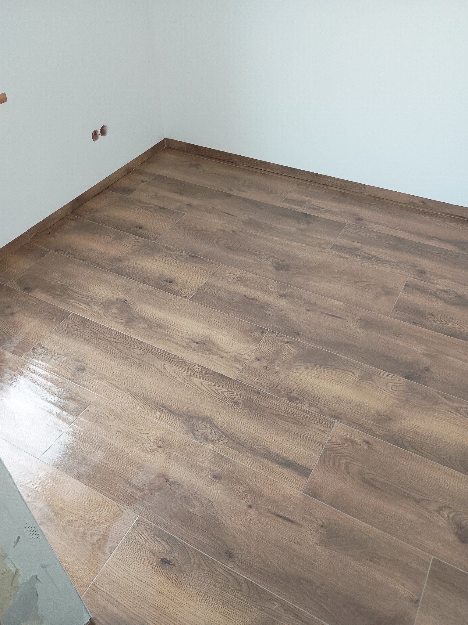 Ukończona podłoga z płytek imitujących drewno w rogu pomieszczenia z białymi ścianami i widocznymi gniazdkami elektrycznymi. Widok z góry, ukośny kadr.