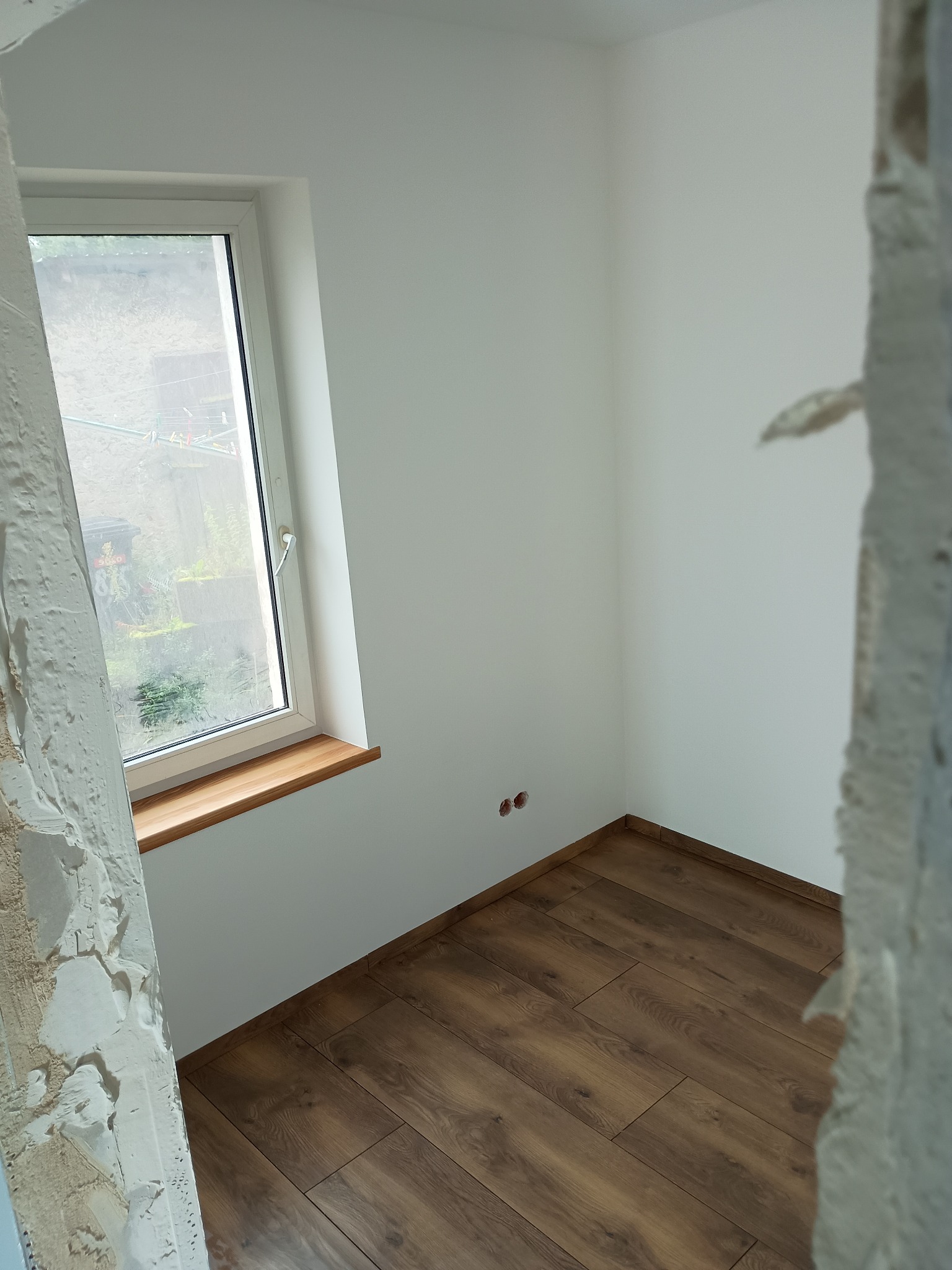 Pokój po remoncie: brązowe panele podłogowe, białe ściany z gniazdkami, okno z drewnianym parapetem. Fragment ściany w stanie surowym po lewej stronie kadru.