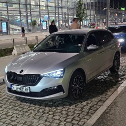 FHU PROPER CARS DAMIAN DUDEK - Srebrna Skoda Scala zaparkowana na brukowanej ulicy przed Terminalem B lotniska w nocy, z włączonymi światłami i widocznymi pasażerami w tle.