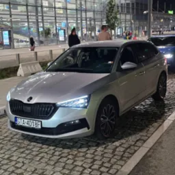 Srebrna Skoda Scala zaparkowana na brukowanej ulicy przed Terminalem B lotniska w nocy, z włączonymi światłami i widocznymi pasażerami w tle.