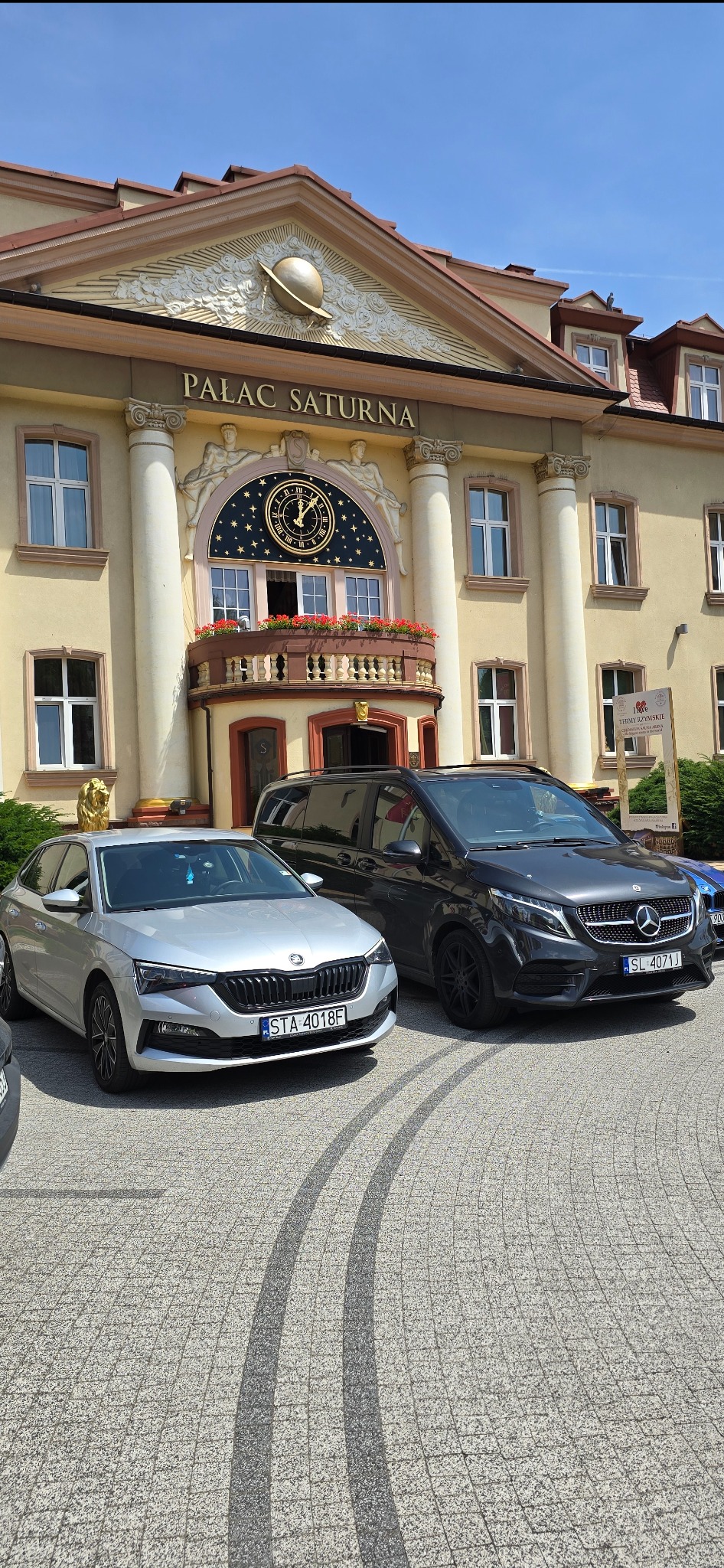 Dwa samochody, Skoda i Mercedes V-klasa, zaparkowane przed budynkiem Pałacu Saturna z charakterystycznym zegarem i balkonem.