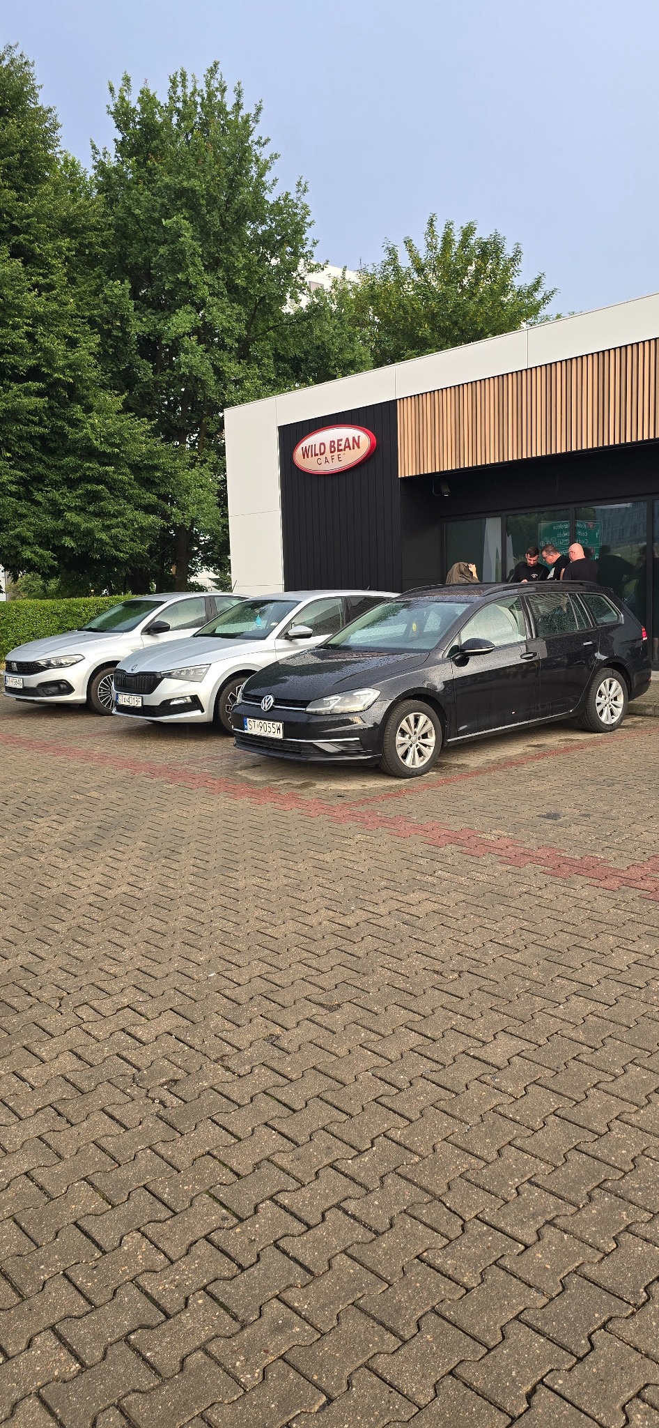 Czarny Volkswagen Golf Variant zaparkowany przed kawiarnią Wild Bean Cafe, obok srebrnego Fiata Tipo i srebrnej Skody Scala. W tle widoczni ludzie przy wejściu do kawiarni.