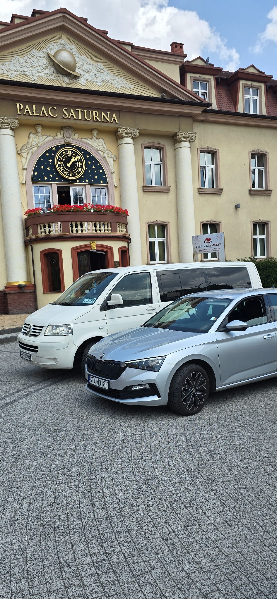 Dwa pojazdy: biały van Volkswagen Transporter i srebrna Skoda Scala, zaparkowane przed budynkiem Pałacu Saturna w Czeladzi.