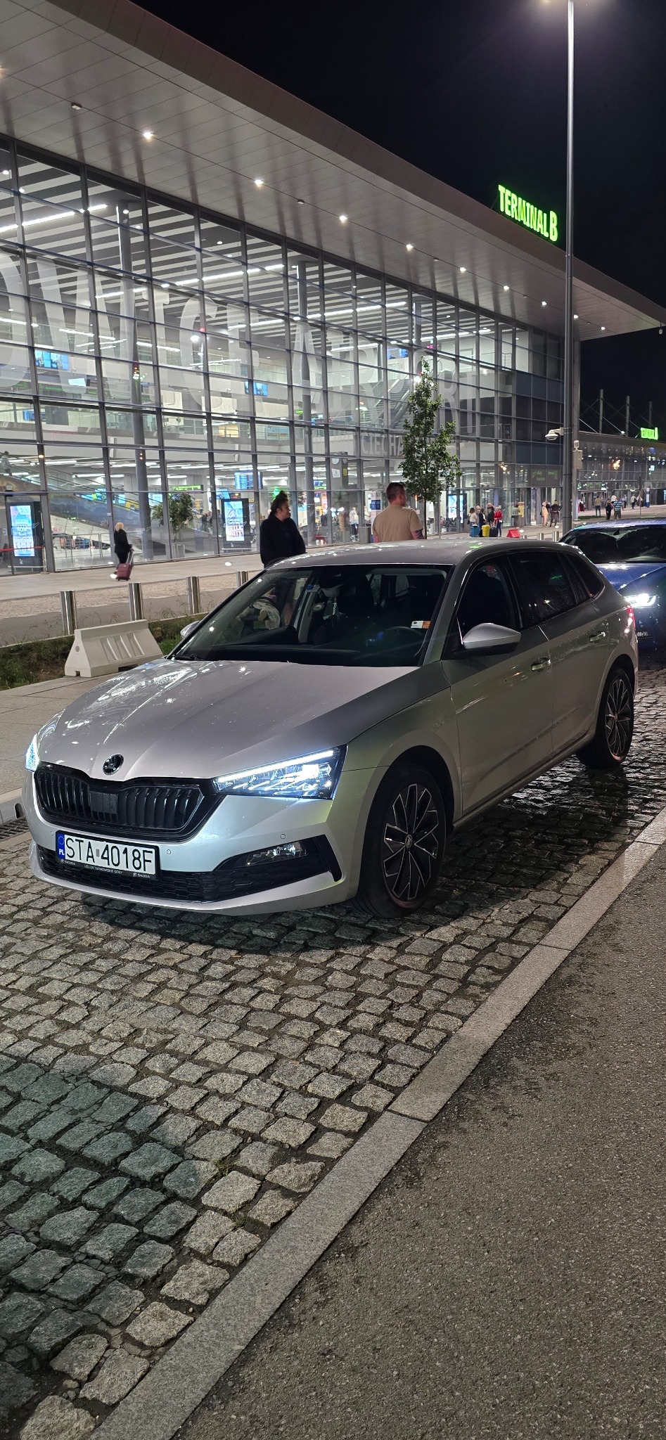 Srebrna Skoda Scala zaparkowana na brukowanej ulicy przed Terminalem B lotniska w nocy, z włączonymi światłami i widocznymi pasażerami w tle.